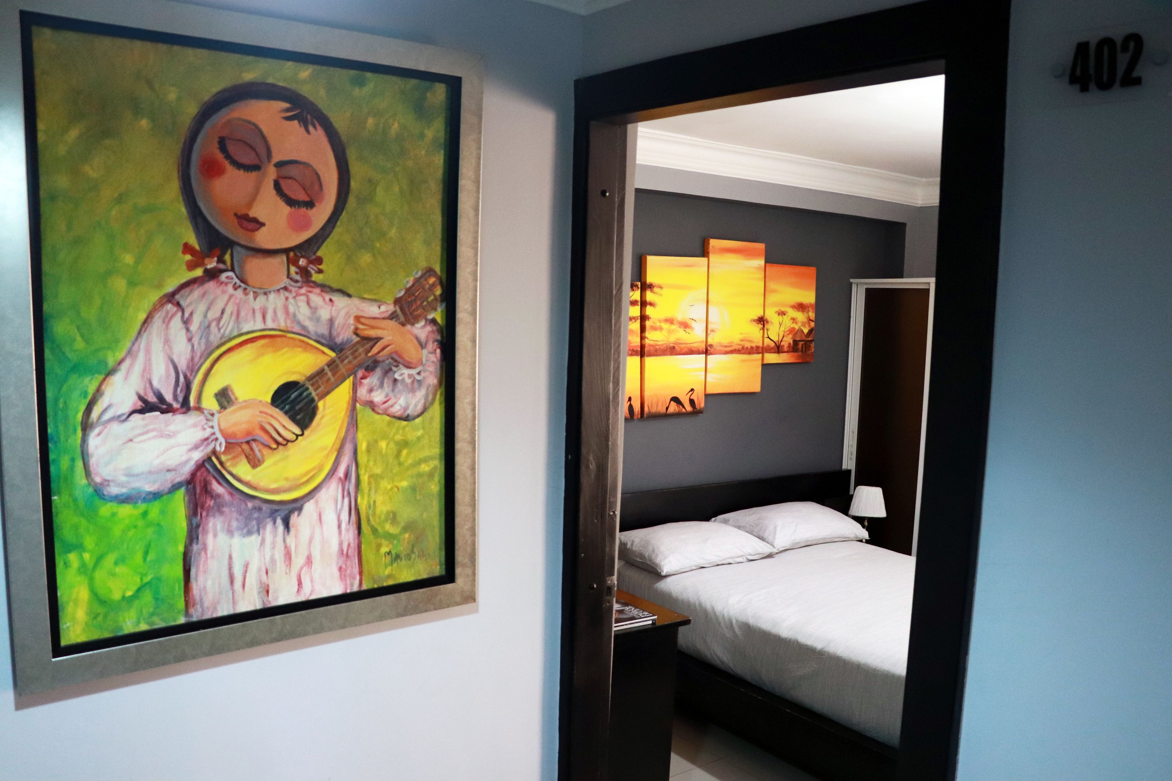 Een hotelkamer met een bed, een nachtkastje en een schilderij van een vrouw die gitaar speelt.
