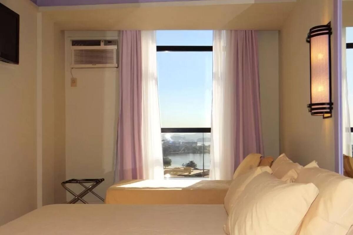 A hotel room with a bed, a window with curtains, an air conditioning unit, and a small table.