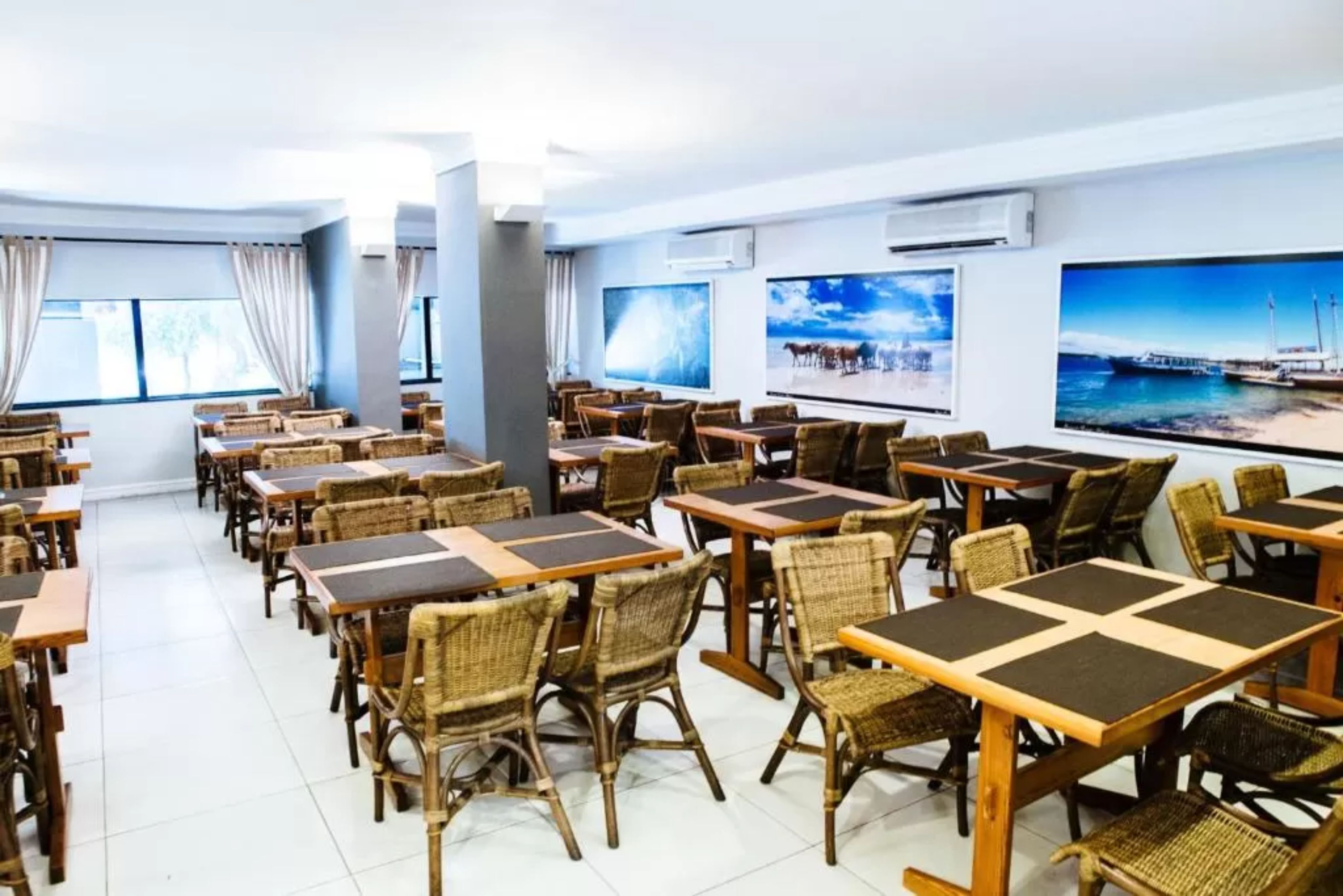 Un comedor con sillas y mesas de mimbre, ventanas amplias con cortinas y obras de arte en las paredes.