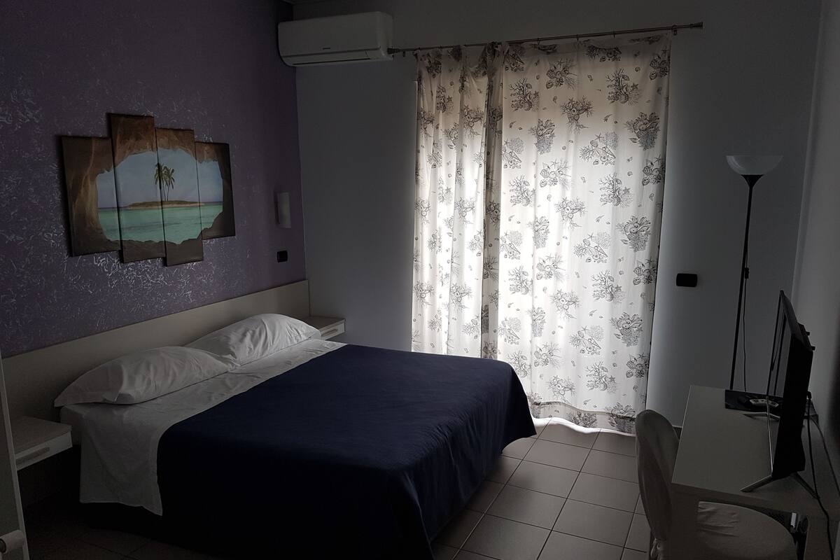 A bedroom with a bed, a nightstand, a desk, a chair, a lamp, and a window with curtains.