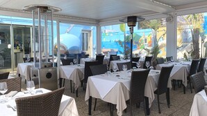 Un área de comedor al aire libre con mesas dispuestas para dos personas, cubierta por un toldo blanco y equipada con calentadores.