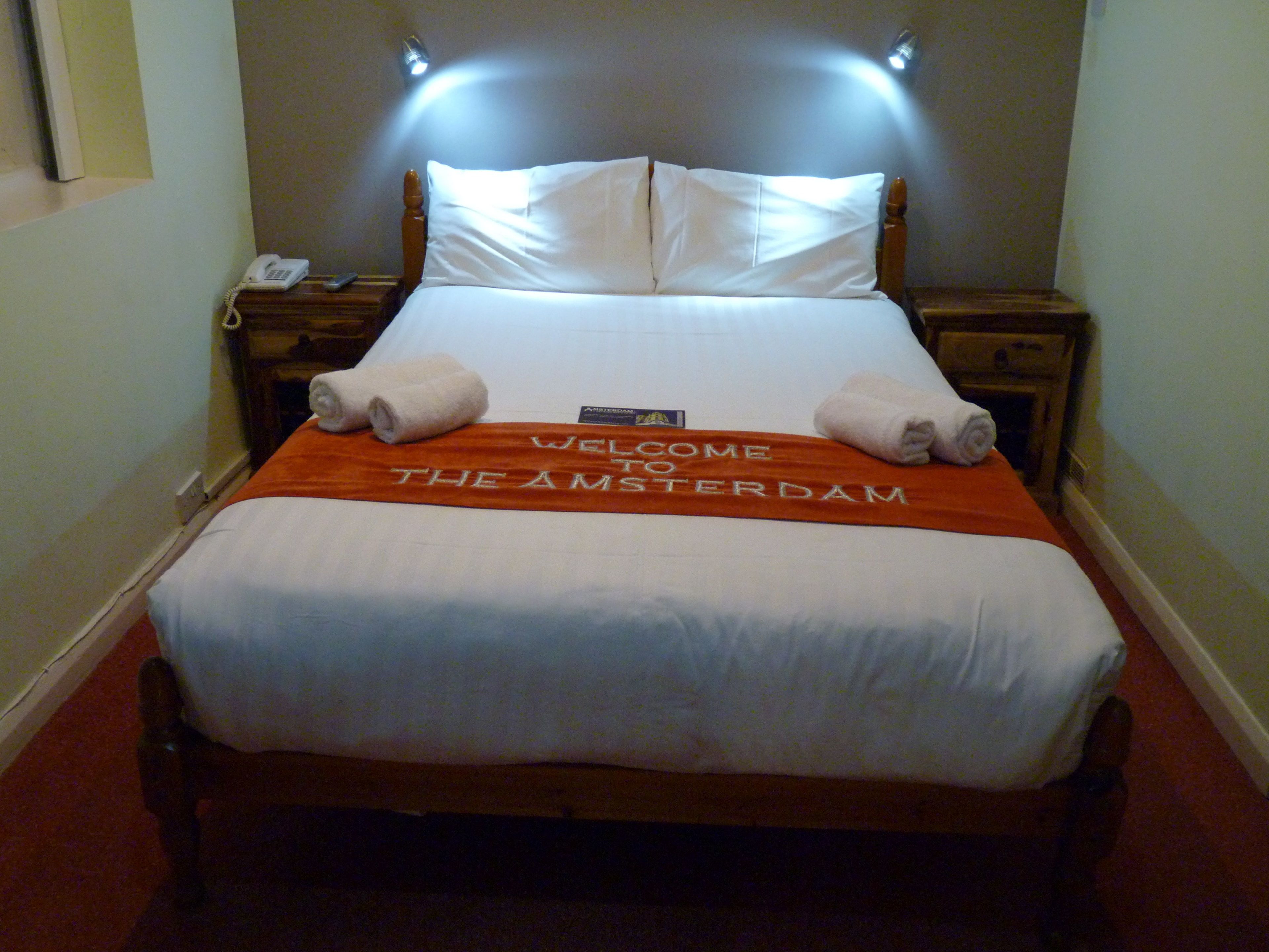Una camera d'albergo con un letto, un comodino, una lampada e un asciugamano di benvenuto.