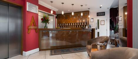 A hotel reception area with a red wall, a reception desk, a seating area with a sofa, and a large mirror.