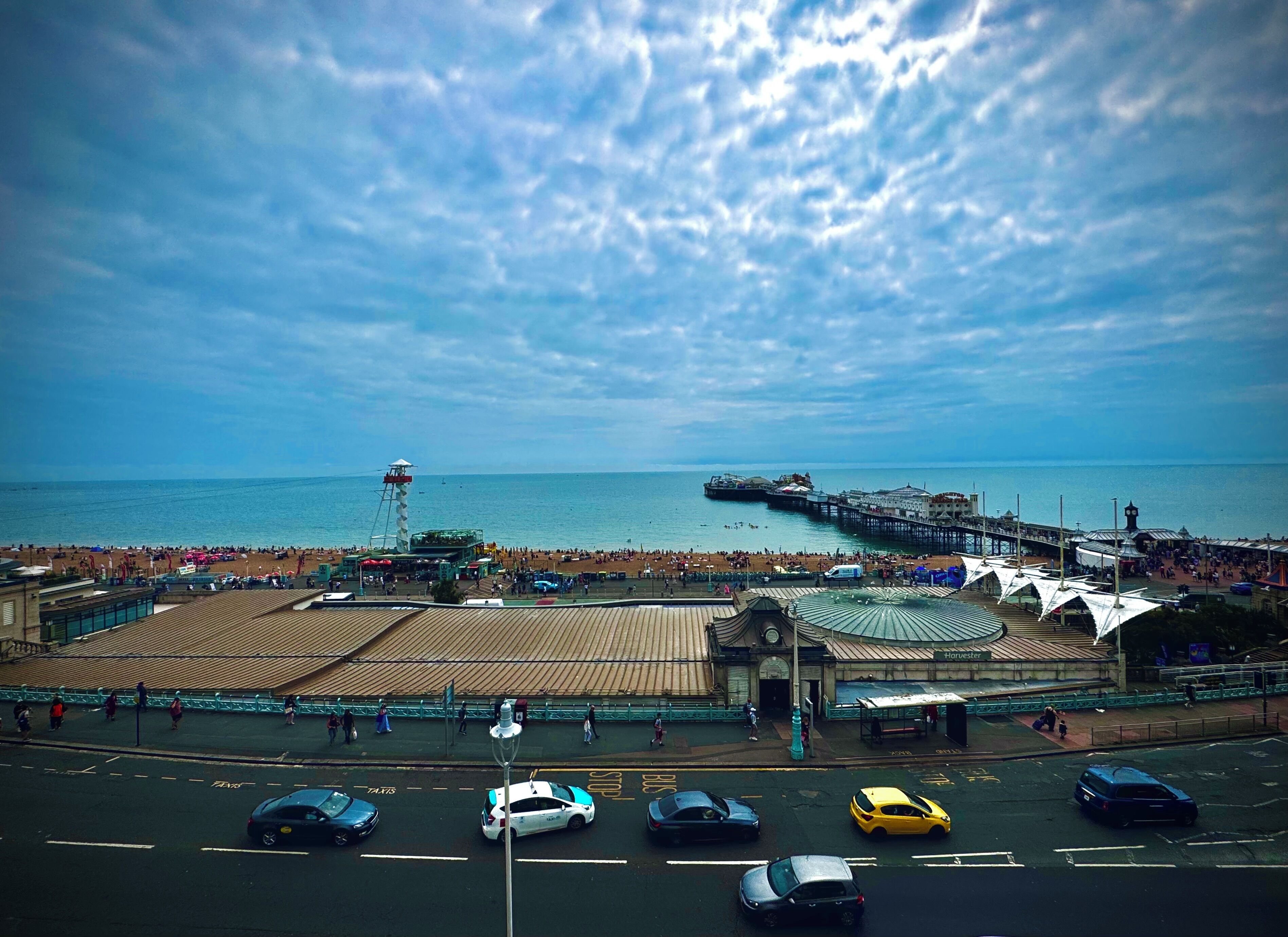 A coastal promenade with a pier extending into the sea, a beach with people, and a parking area with cars.