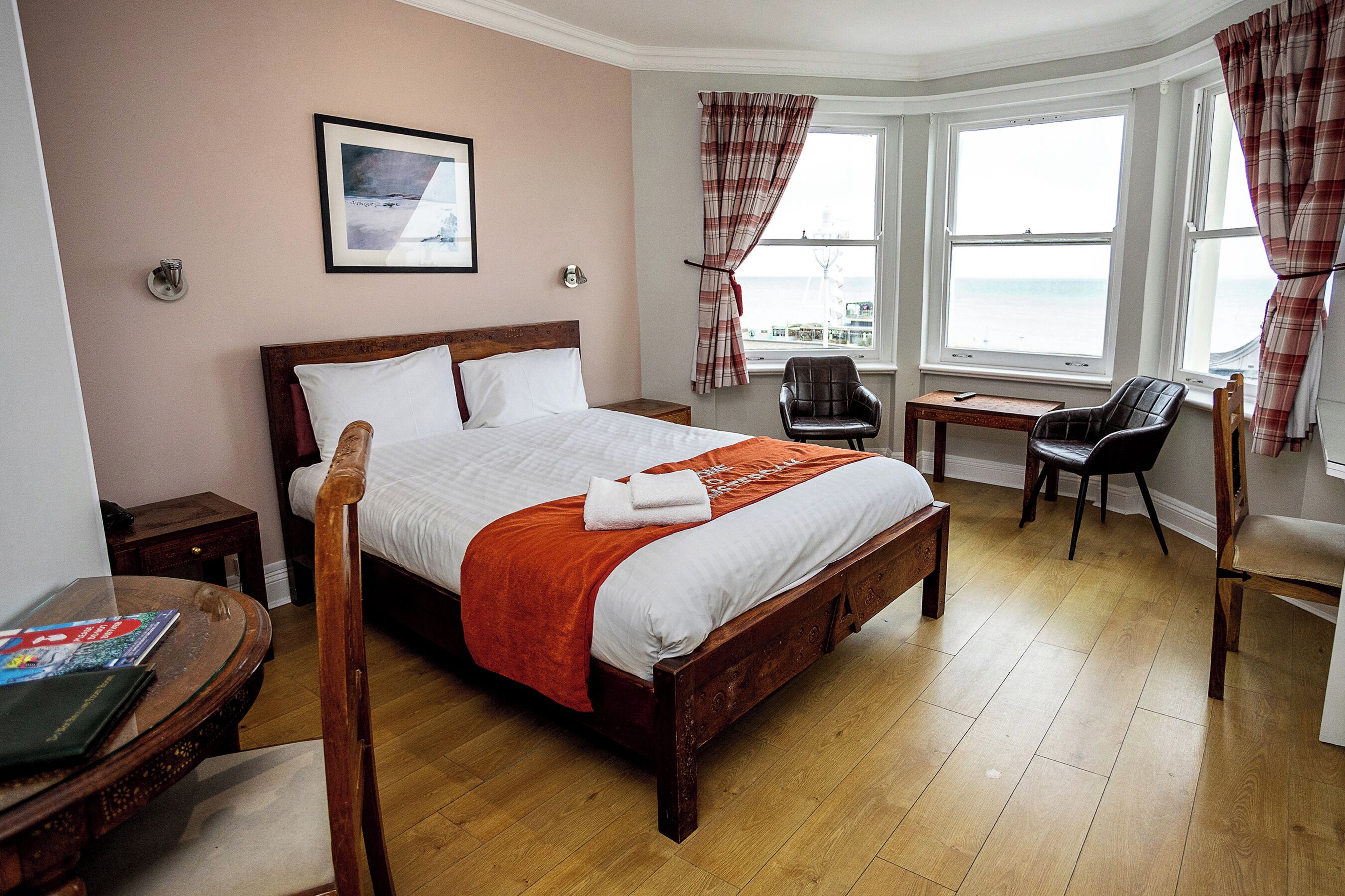 A neatly arranged bedroom with a large bed, a wooden chair, a small table, and a view of the sea through the window.