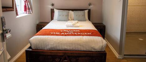 Una cama bien tendida con un letrero que dice "WELCOME TO THE AMSTERDAM", un cabecero de madera y una mesita de noche con una lámpara.