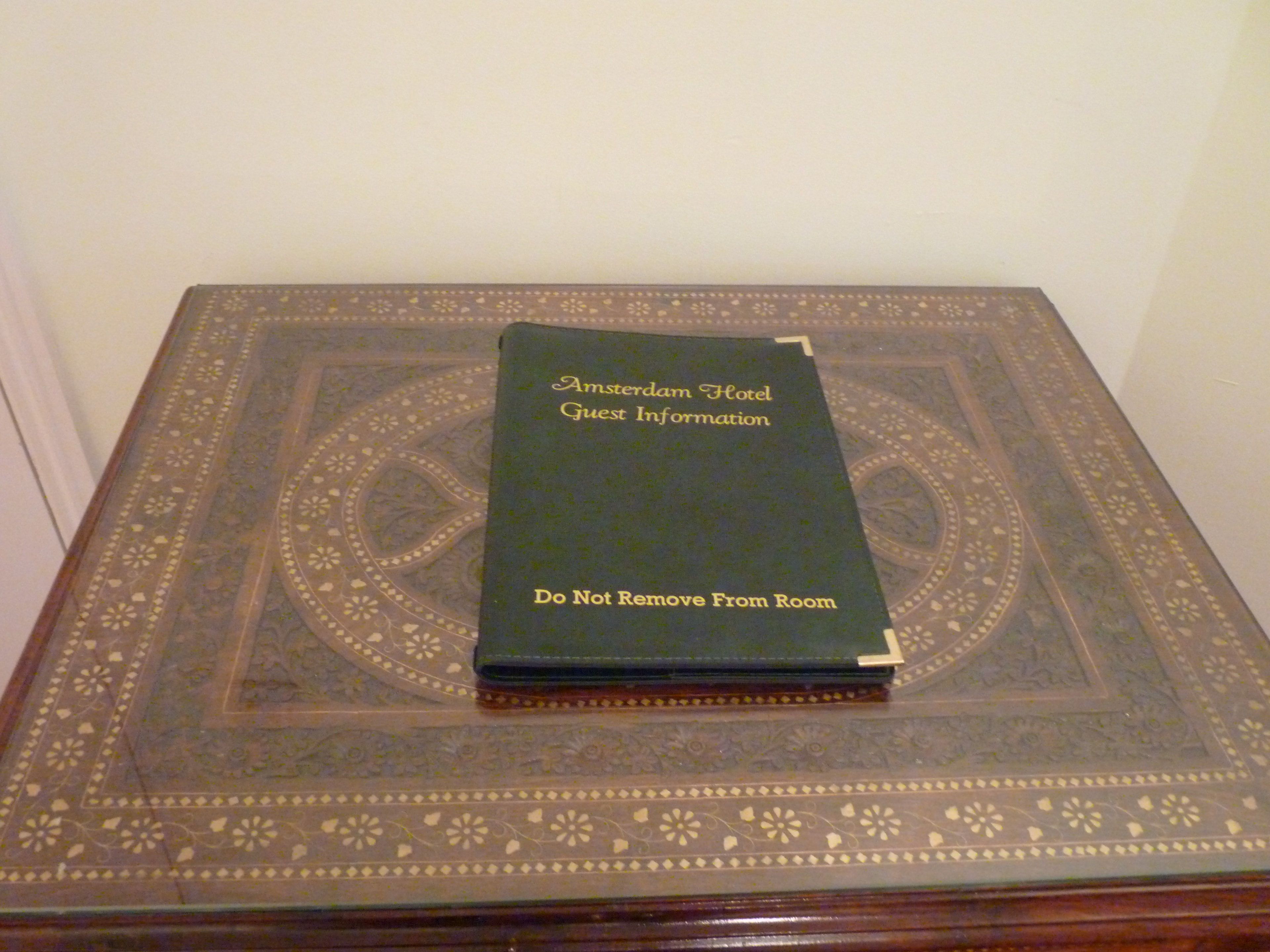 A book titled "Amsterdam Hotel Guest Information" with a warning "Do Not Remove From Room" on a patterned table.