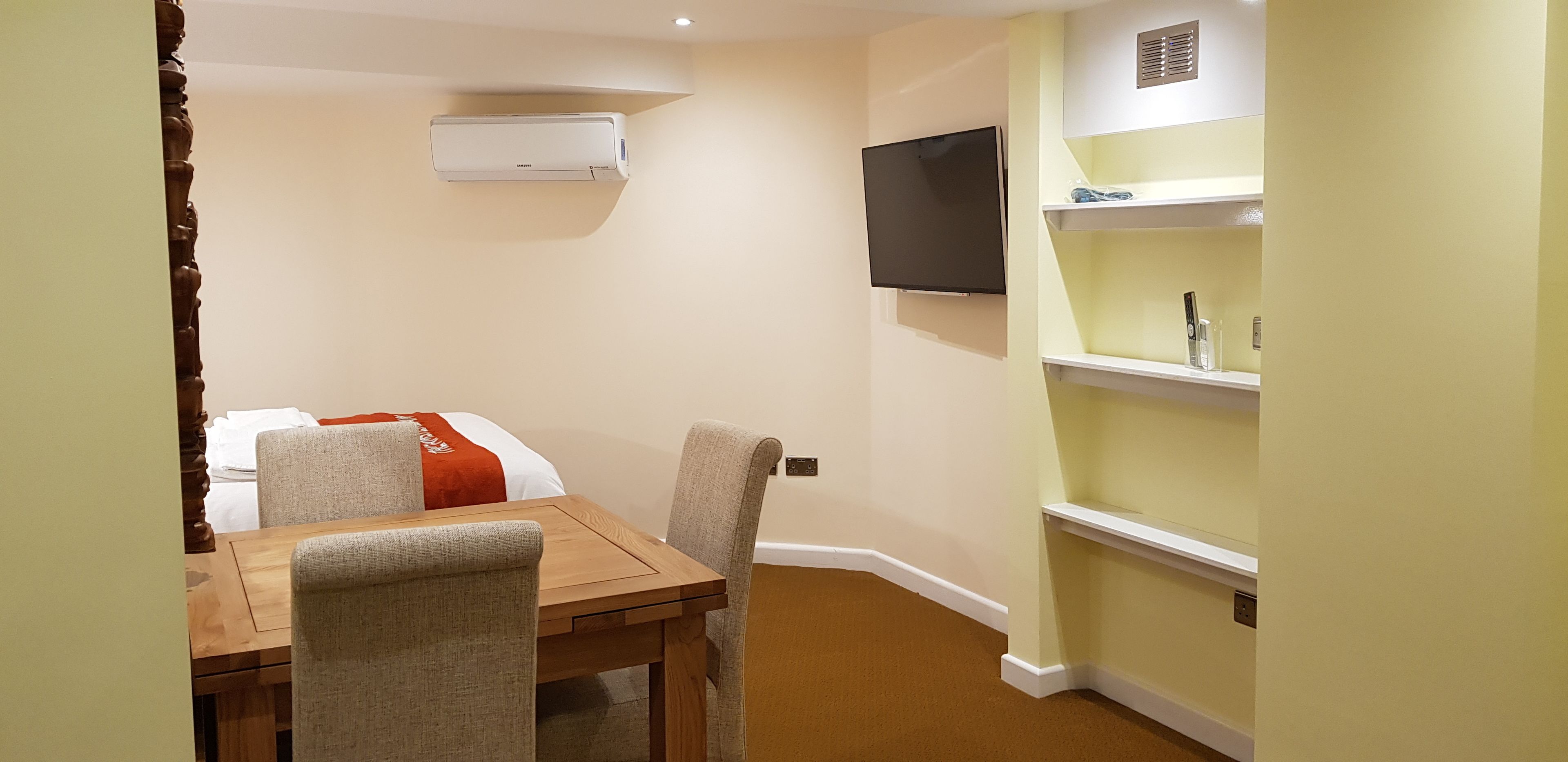 A hotel room with a wooden table, two chairs, a bed with a red blanket, an air conditioner, a TV mounted on the wall, and a small shelf with toiletries.