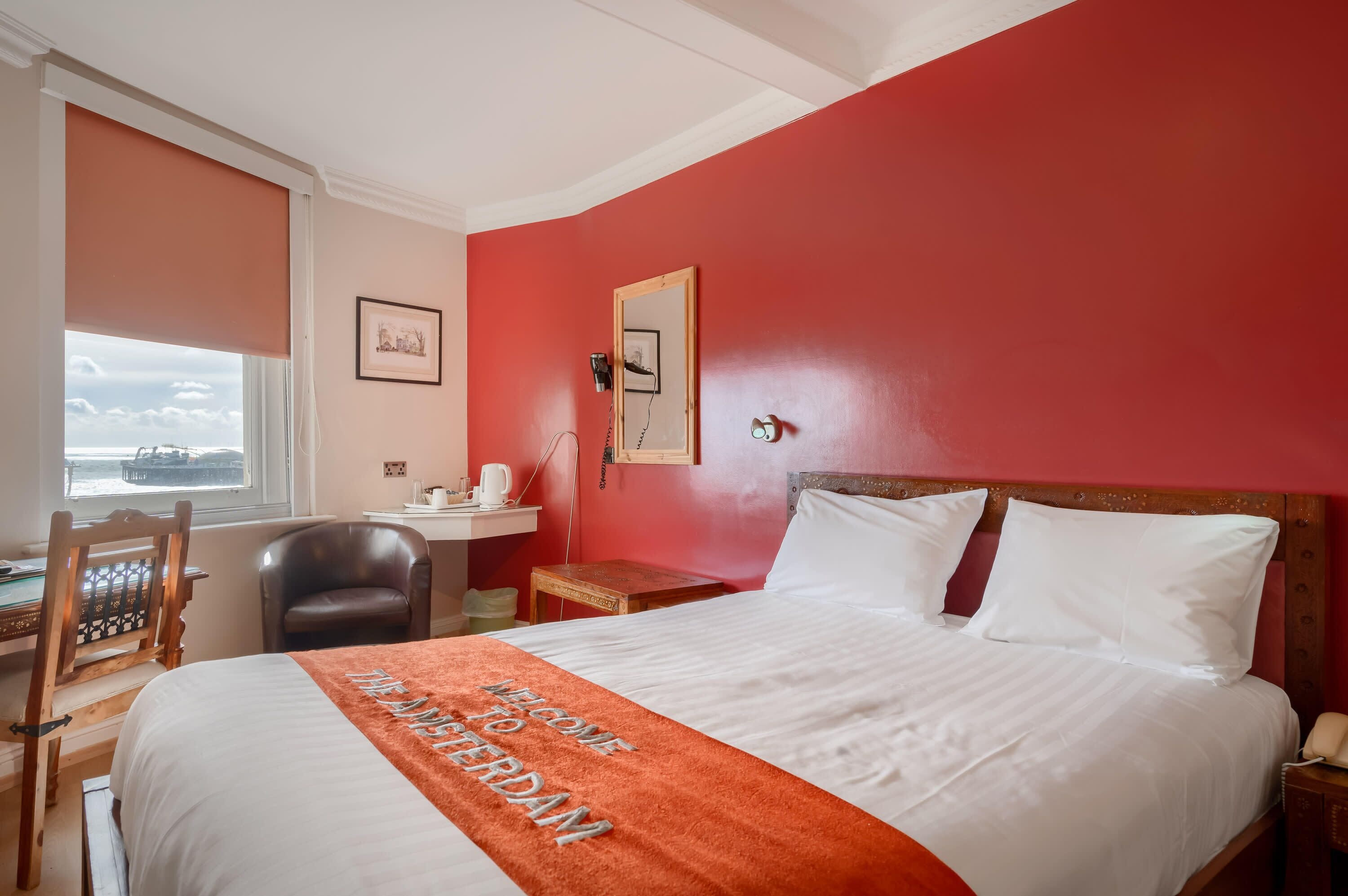 A hotel room with a bed, a desk, a chair, a small table, a telephone, and a window with a view of the sea.