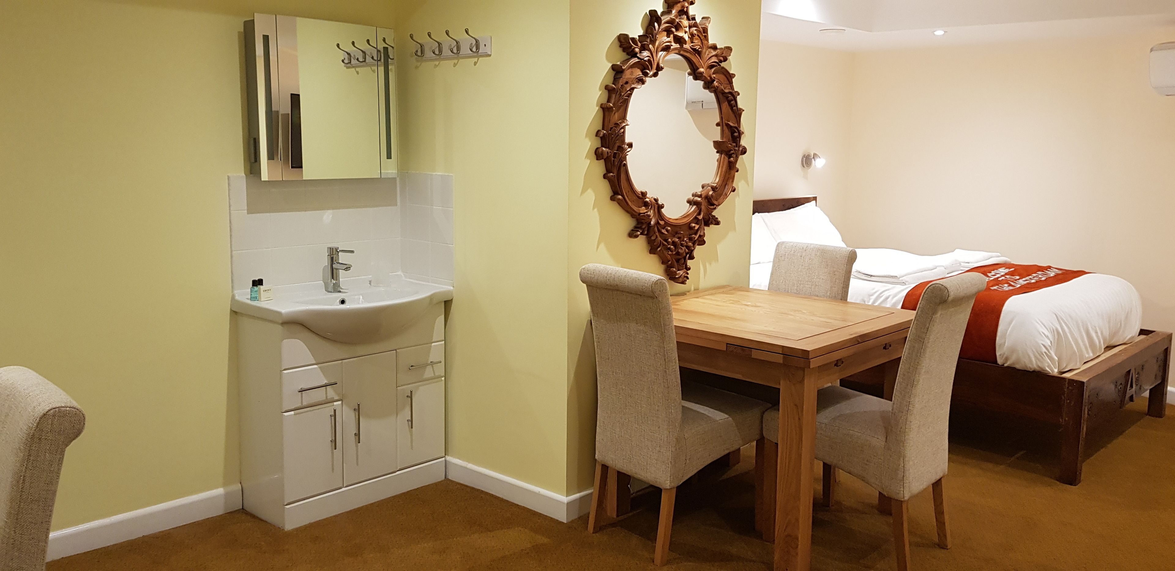 A hotel room with a wooden dining table, two chairs, a bed with a red blanket, a large ornate mirror, and a white sink with cabinets.