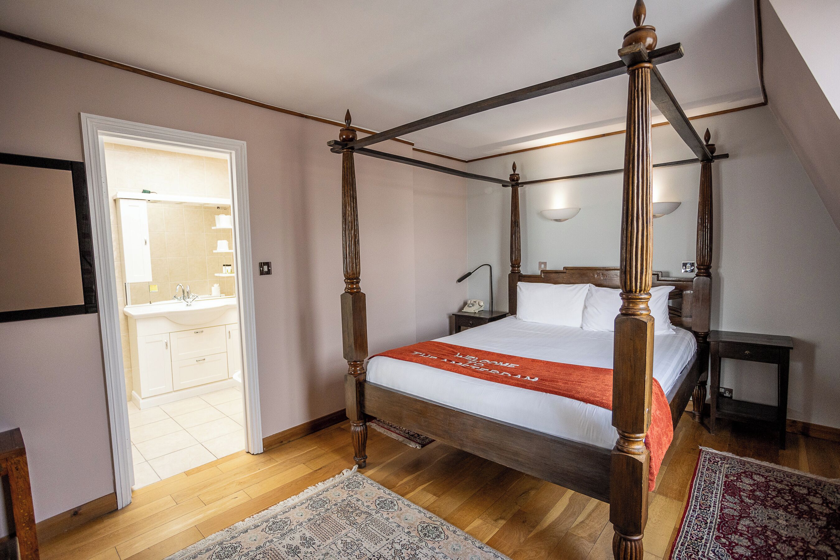 A four-poster bed with a wooden frame and white bedding, a bedside table with a lamp, and a bathroom with a sink and mirror.