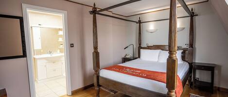 A four-poster bed with a wooden frame and white bedding, a bedside table with a lamp, and a bathroom with a sink and mirror.