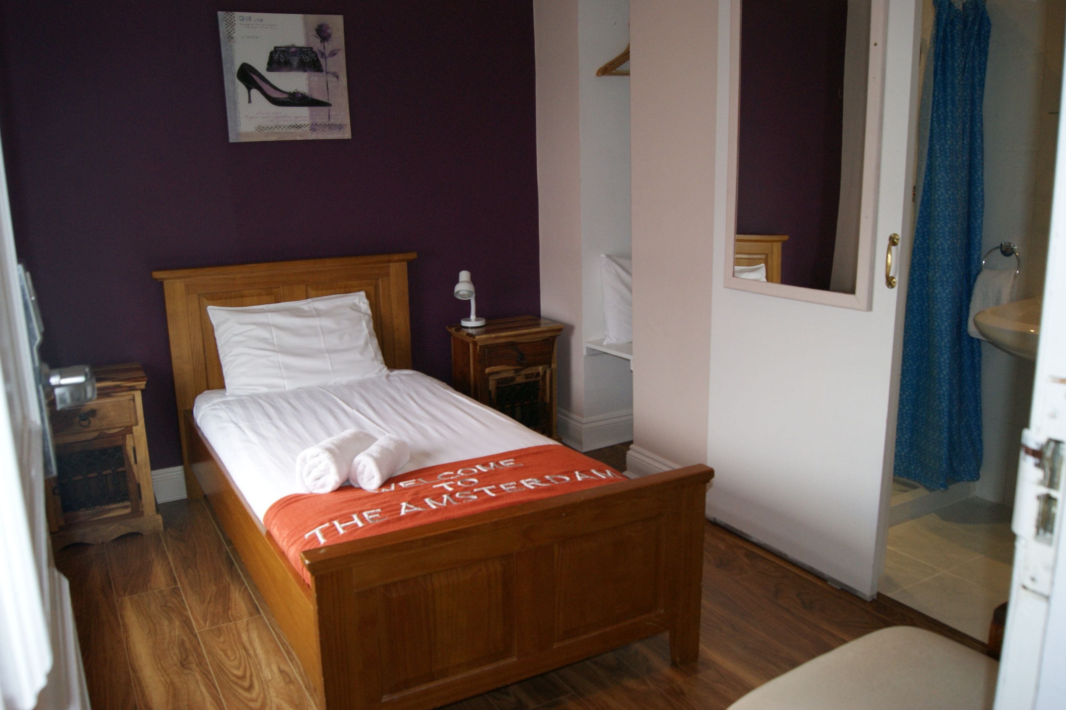 Een netjes opgemaakt bed met een witte deken en kussens, een houten hoofdbord, een nachtkastje met een lamp, een spiegel aan de muur, en een badkamer met een wastafel en douchegordijn.