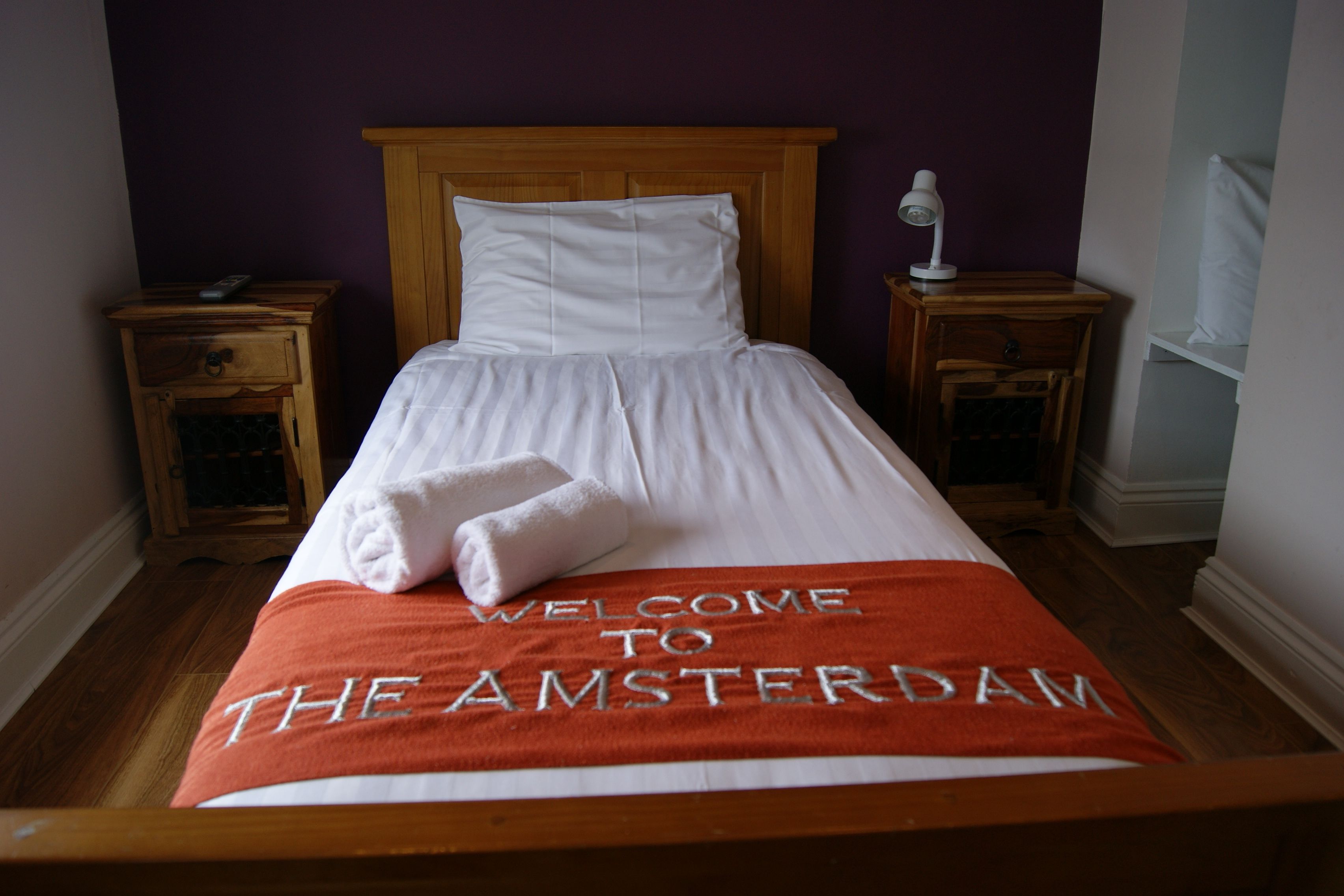 A neatly made bed with a "Welcome to The Amsterdam" blanket, wooden headboard, two bedside tables with lamps, and a purple wall.