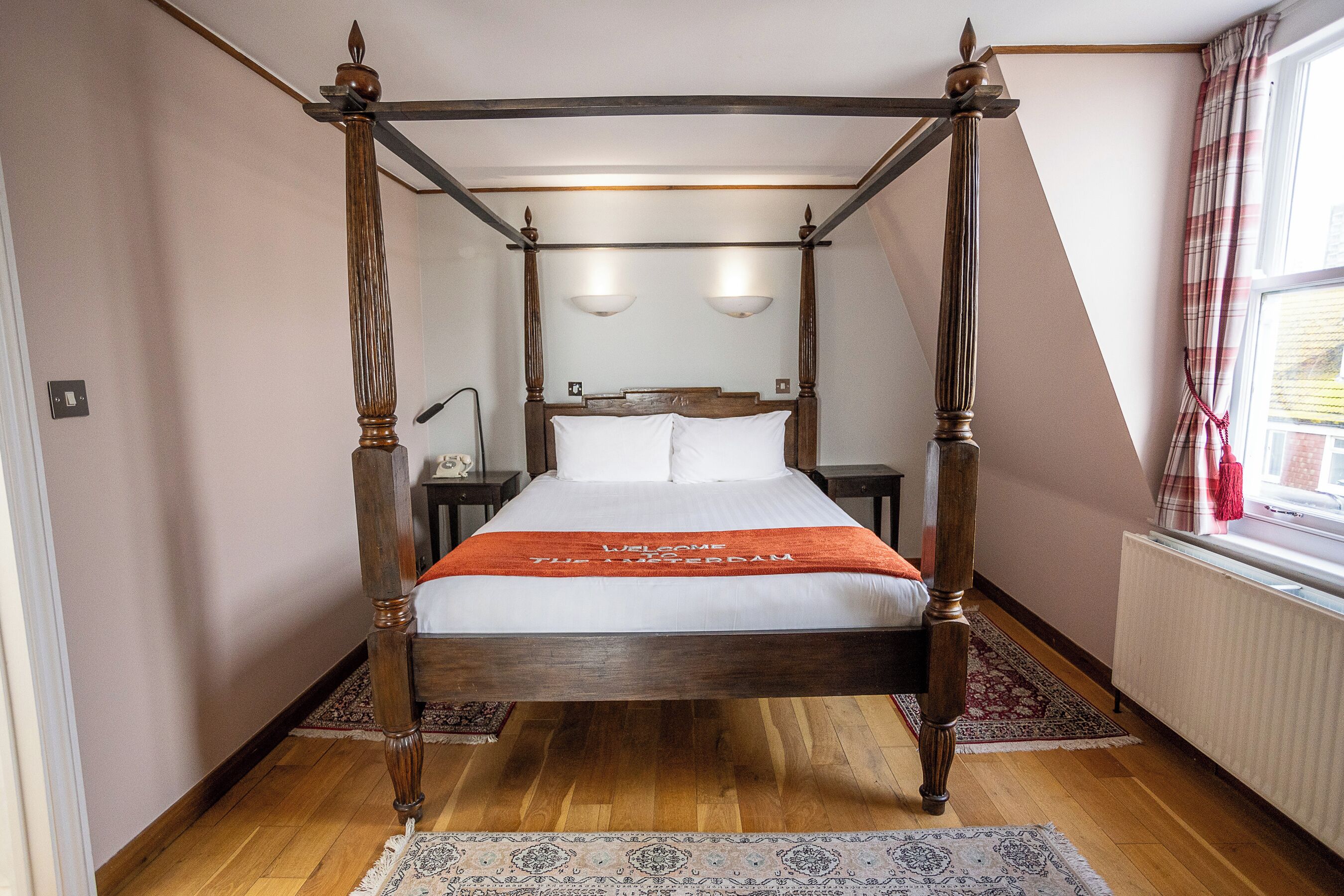 Una cama con dosel, un edredón que dice “Bienvenido al Hotel”, suelo de madera y una ventana con cortinas.