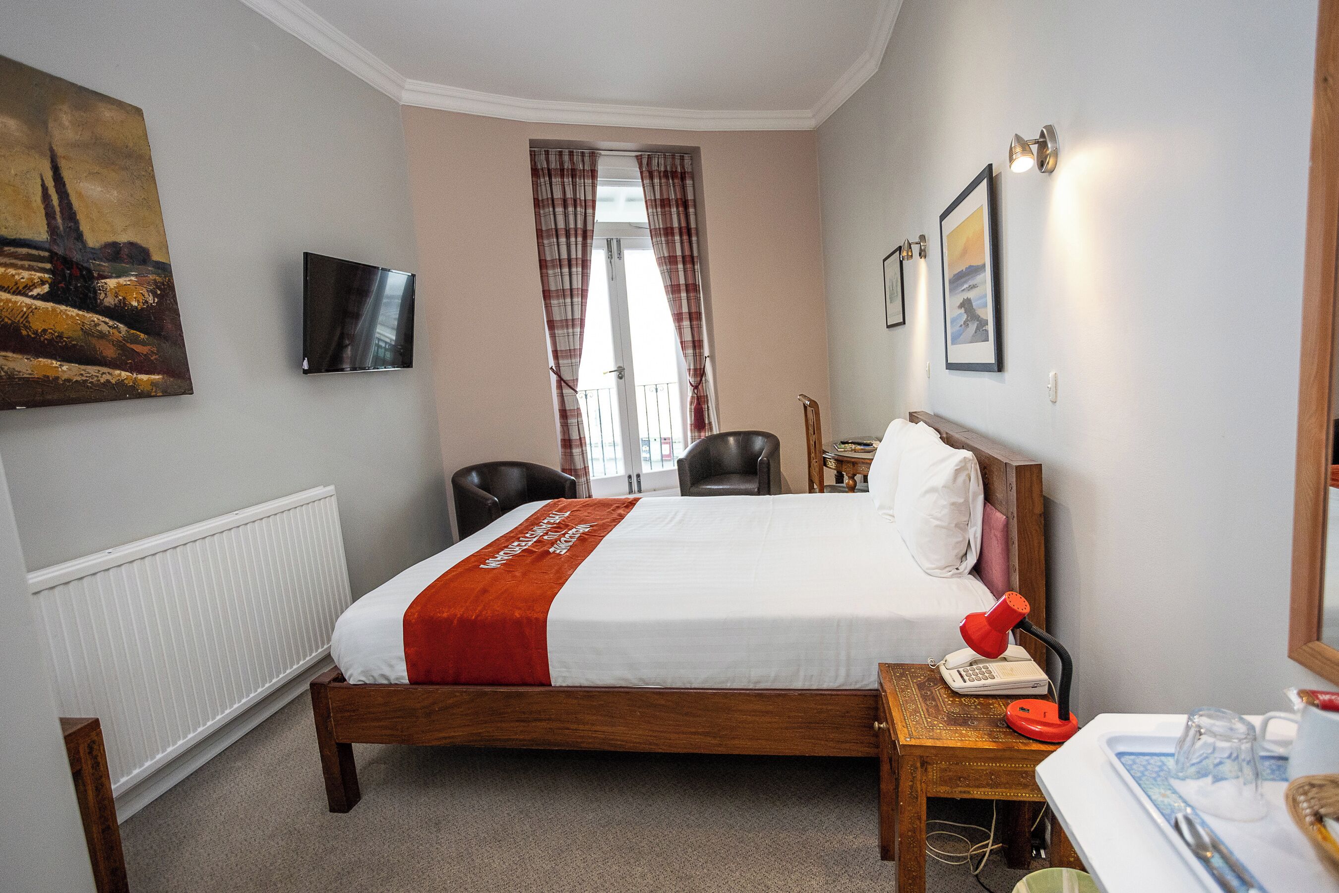 A hotel room with a bed, a desk with a telephone, a chair, a television mounted on the wall, and a window with curtains.