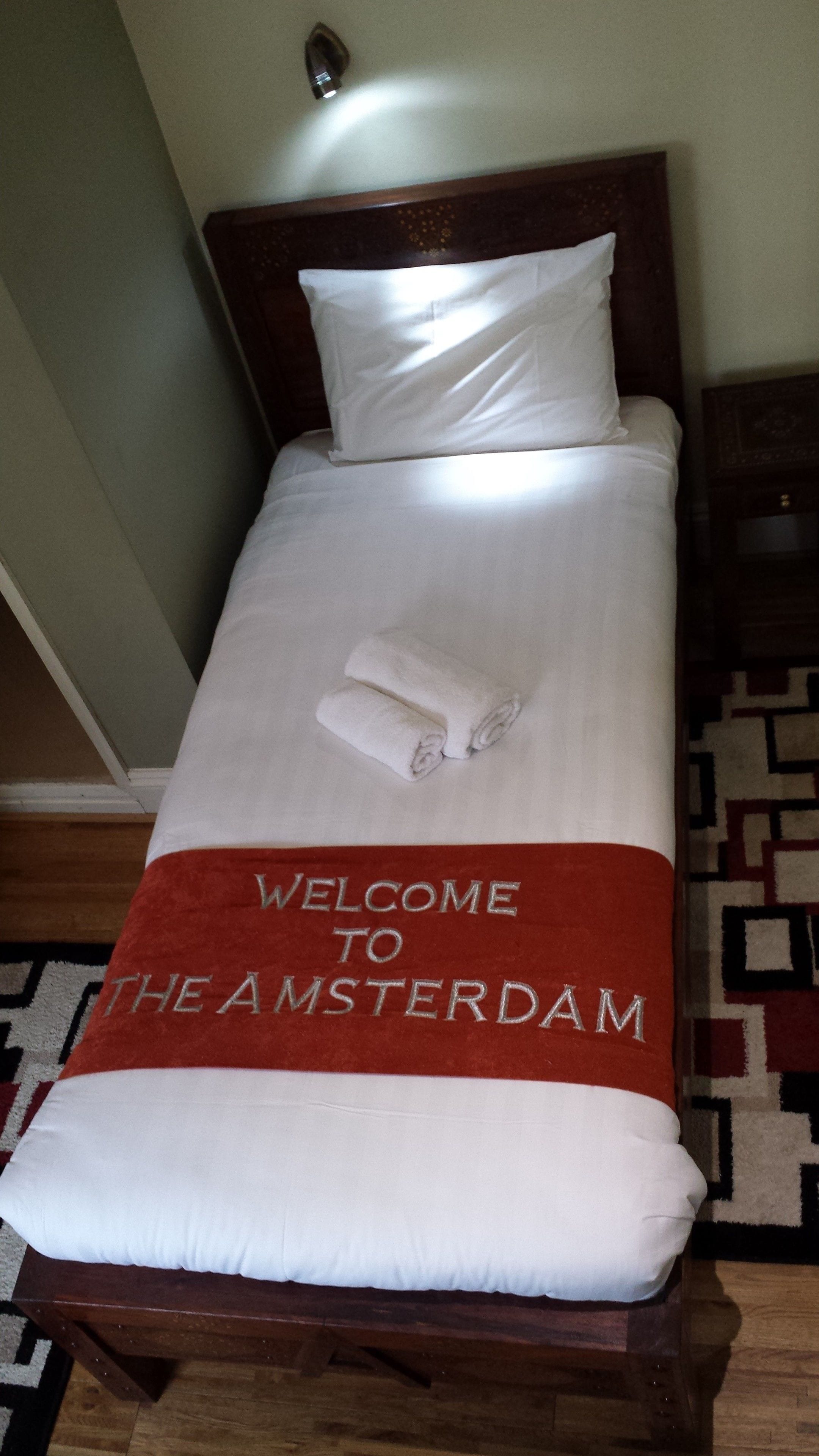 Una cama bien hecha con un edredón blanco y almohadas, una manta roja con el texto "WELCOME TO THE AMSTERDAM" y un par de toallas enrolladas.