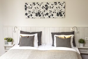 A neatly made bed with grey and white bedding, a floral wall hanging, and two bedside tables with small plants. - Guesthouse Eleven Hotel (Arvika)