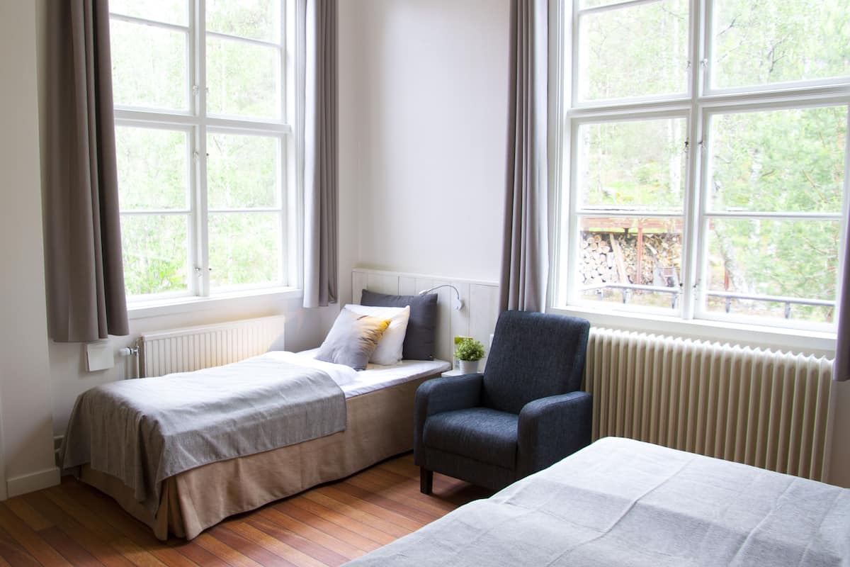 A room with two beds, a chair, a radiator, and large windows with curtains.