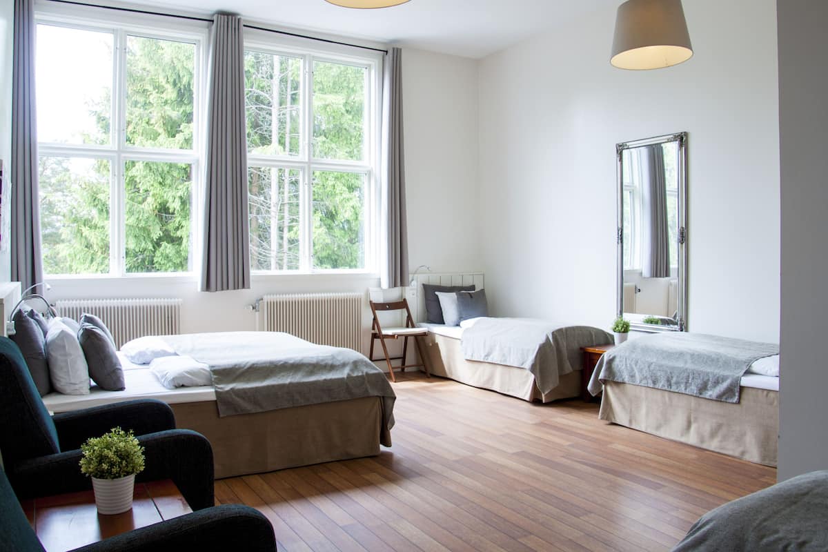 A modern hotel room with two single beds, a large window with curtains, a wooden floor, a chair, a small table, and a potted plant.