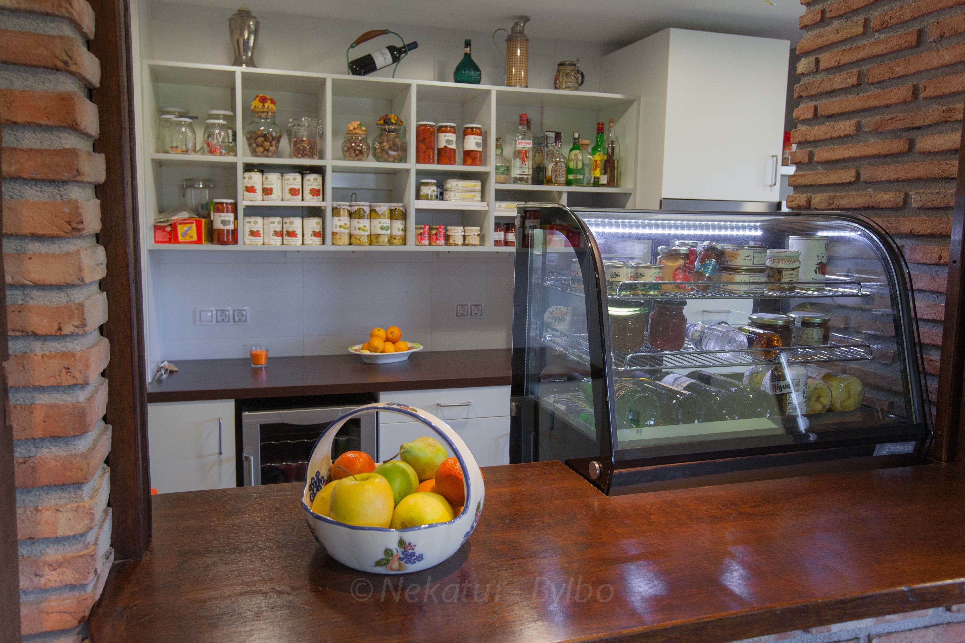 A kitchen with a display case containing various food items, a bowl of fruit on the counter, and shelves stocked with jars and bottles.