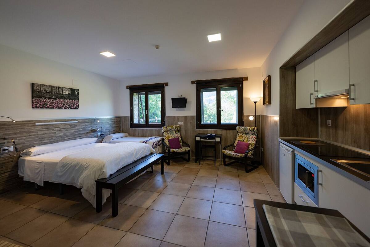 A bedroom with two beds, a desk, a chair, a TV, and a kitchenette.