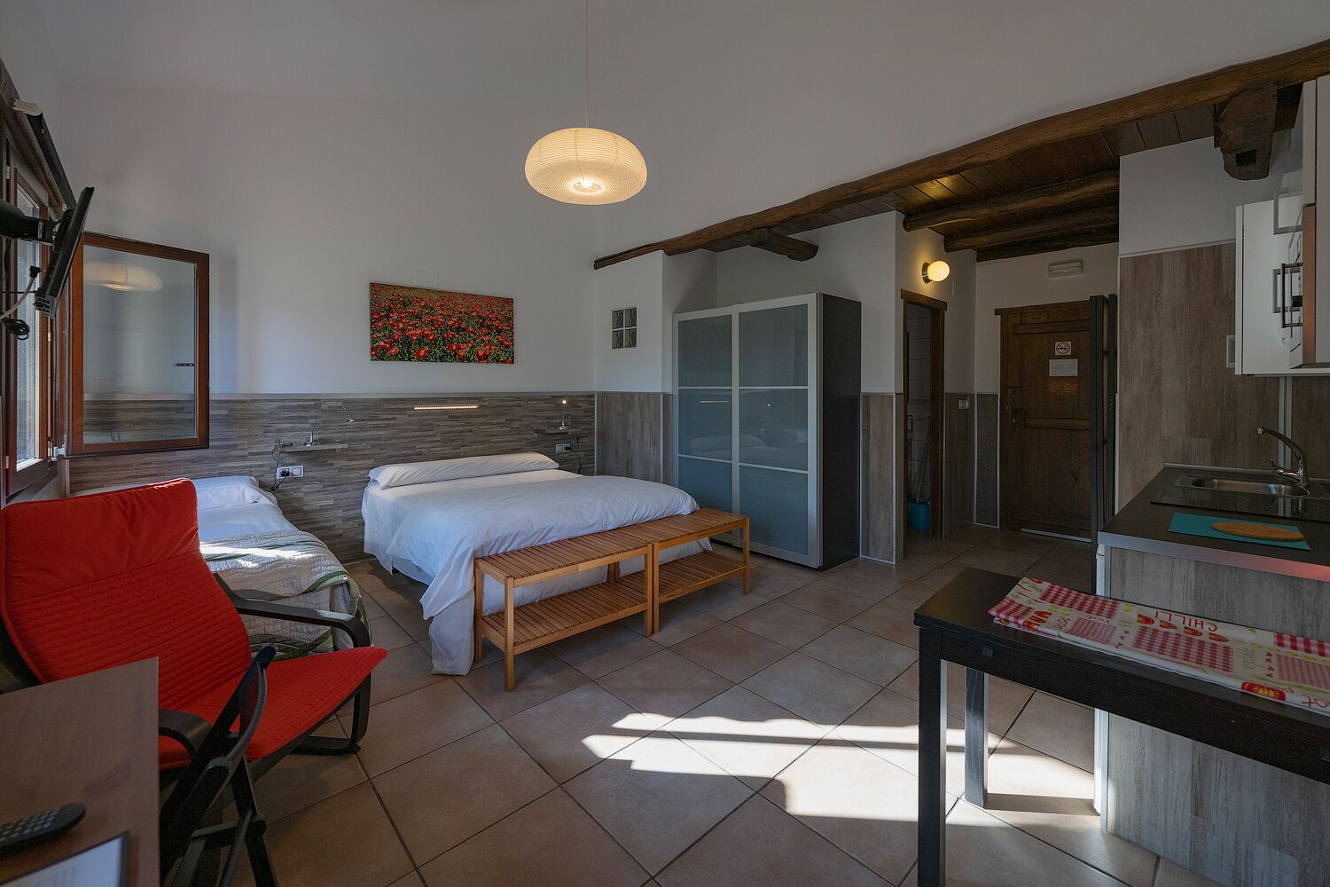 Quarto moderno com cama, um banco de madeira, uma cadeira vermelha, uma escrivaninha e uma área de cozinha com pia.