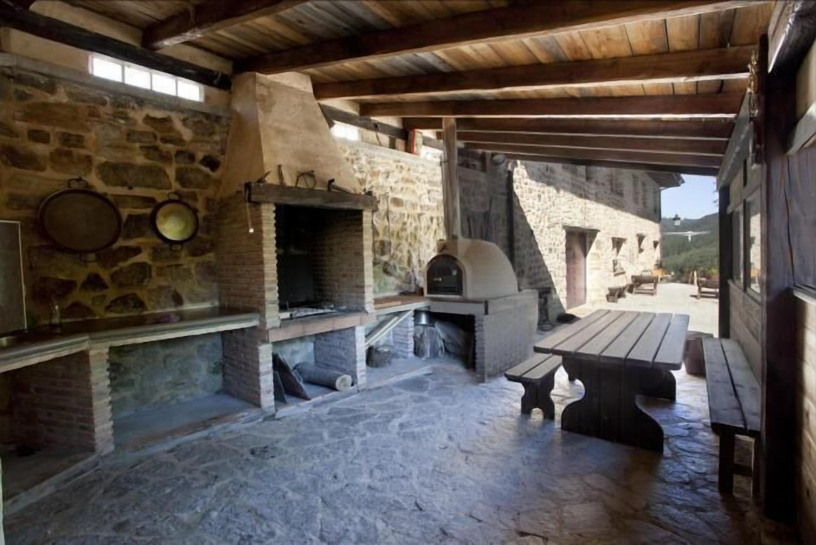 Una cucina esterna in pietra con forno a legna, un tavolo di legno e panche.