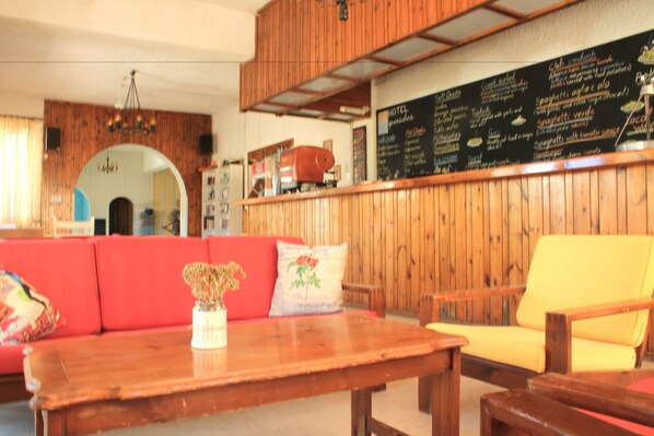 A wooden interior with a red sofa, yellow armchair, and a wooden table with a vase. A chalkboard menu is visible on the wall.