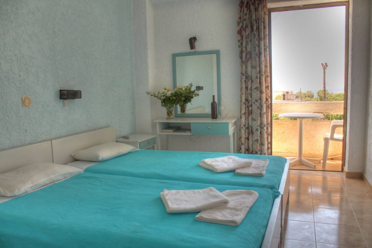 A hotel room with a bed, a nightstand, a mirror, a vase with flowers, and a view of the outside.