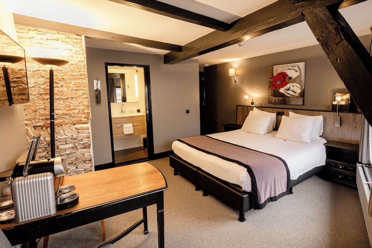 Une chambre avec un lit, une table de chevet et un miroir. On y trouve un banc en bois et une petite table avec une bouilloire et une tasse. La pièce est dotée d’un mur en pierre et d’une poutre en bois au plafond.