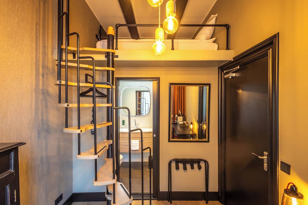 Une chambre d’hôtel compacte avec un lit superposé, un escalier en colimaçon en bois, un miroir et une porte.