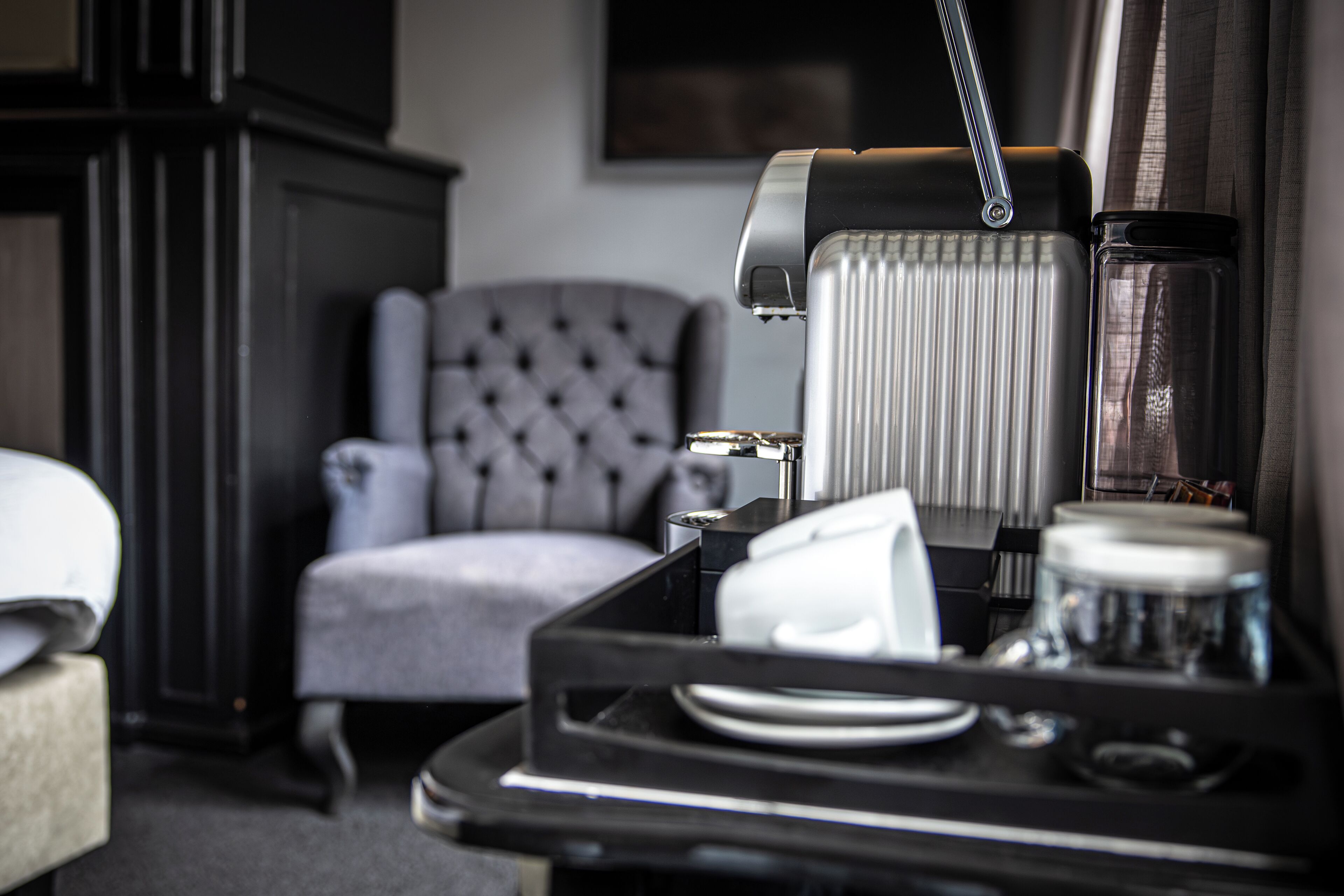 Una habitación de hotel con un carrito de comidas, un sillón gris con capitoné y un radiador.