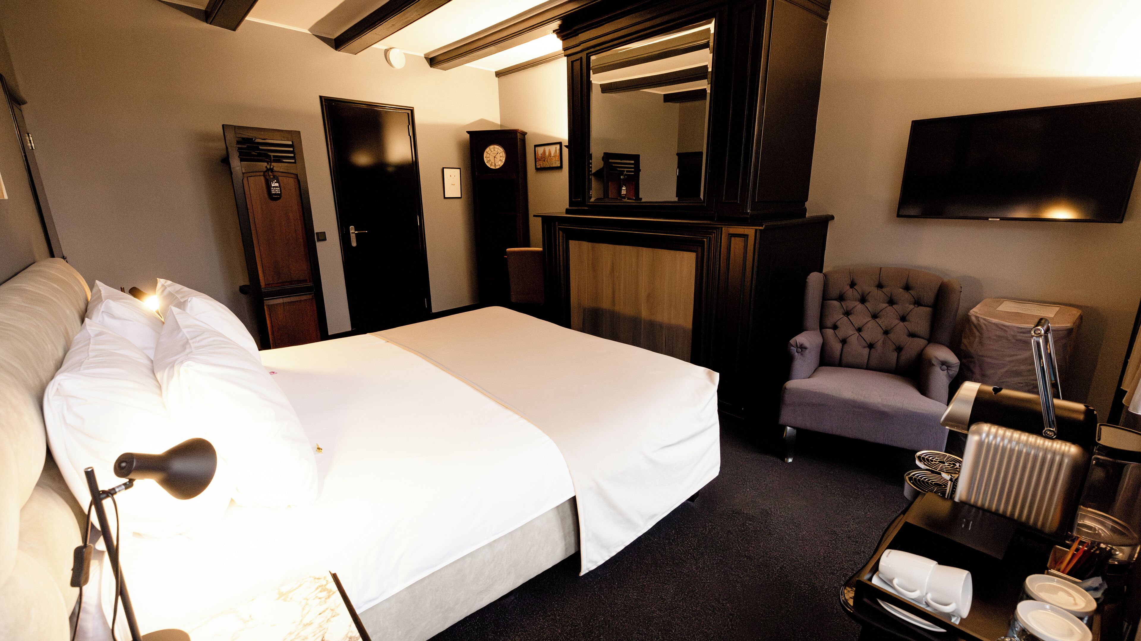 A hotel room with a bed, a chair, a desk with a lamp, a clock, and a television on the wall.