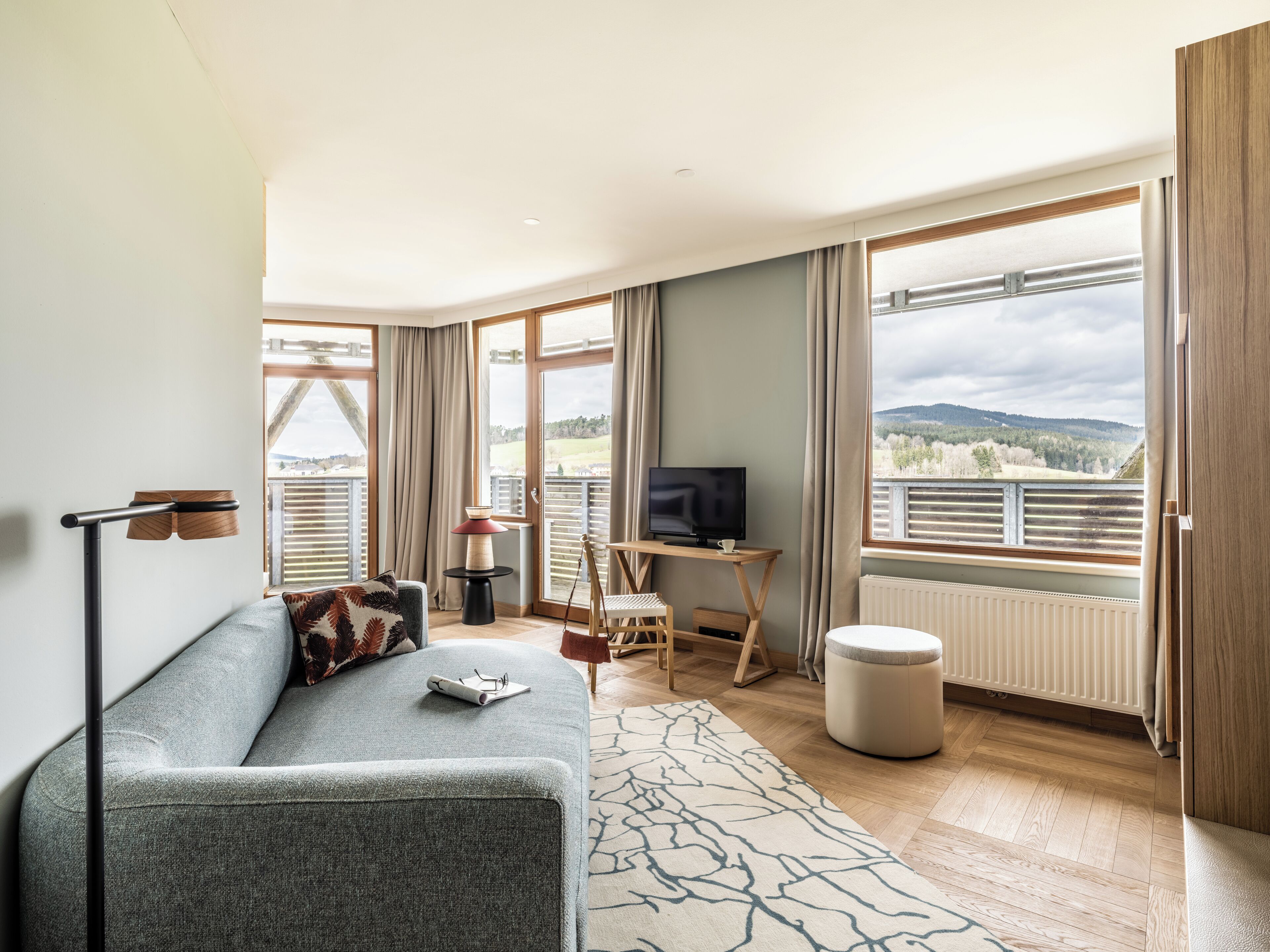 Una sala de estar moderna con un sofá gris, un televisor sobre un mueble de madera, un ventanal con vistas al campo y un piso de madera.