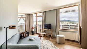Una sala de estar moderna con un sofá gris, un televisor sobre un mueble de madera, un ventanal con vistas al campo y un piso de madera.