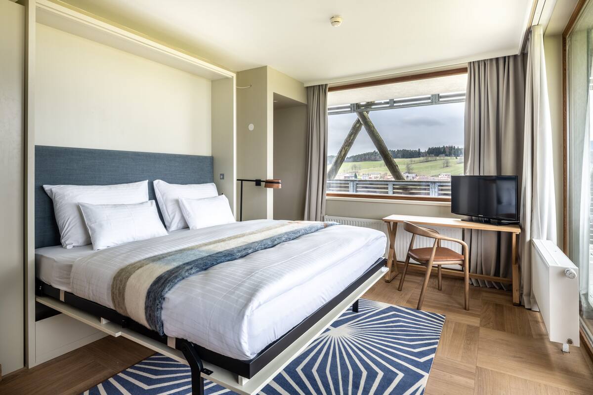 Hotelkamer met een bed, bureau, stoel, televisie en raam met uitzicht op een brug en landschap.