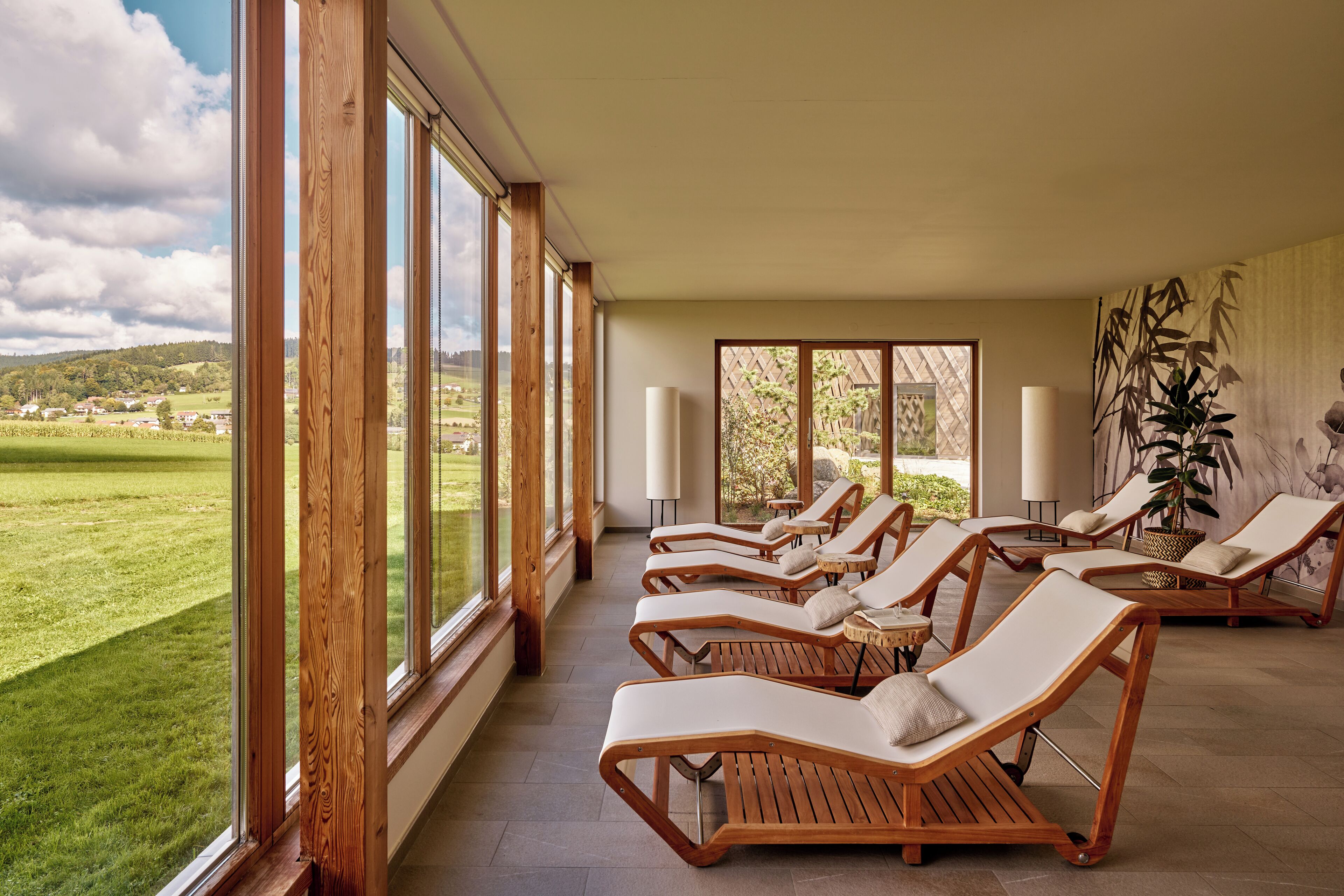 A sunlit room with large windows, wooden lounge chairs, and a view of a green landscape.