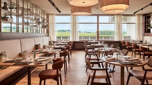 A restaurant with a long dining area, wooden tables, and chairs. There are large windows with a view of the countryside.
