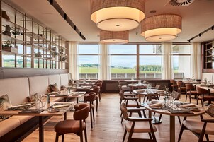 Un restaurante con un amplio comedor, mesas y sillas de madera. Se ven grandes ventanales con vistas al campo.