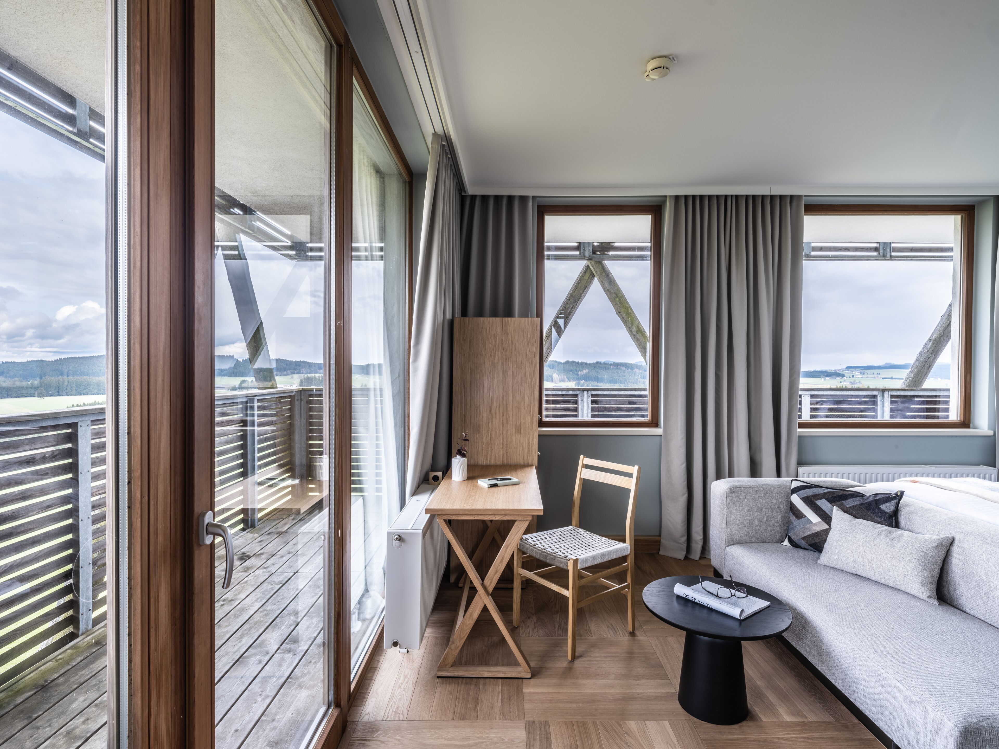 Ein modernes Zimmer mit Holzboden, einem grauen Sofa, einem Holztisch und einem Stuhl. Große Fenster mit Vorhängen, eine Heizung sowie Blick auf eine Brücke und eine Landschaft sind zu sehen.