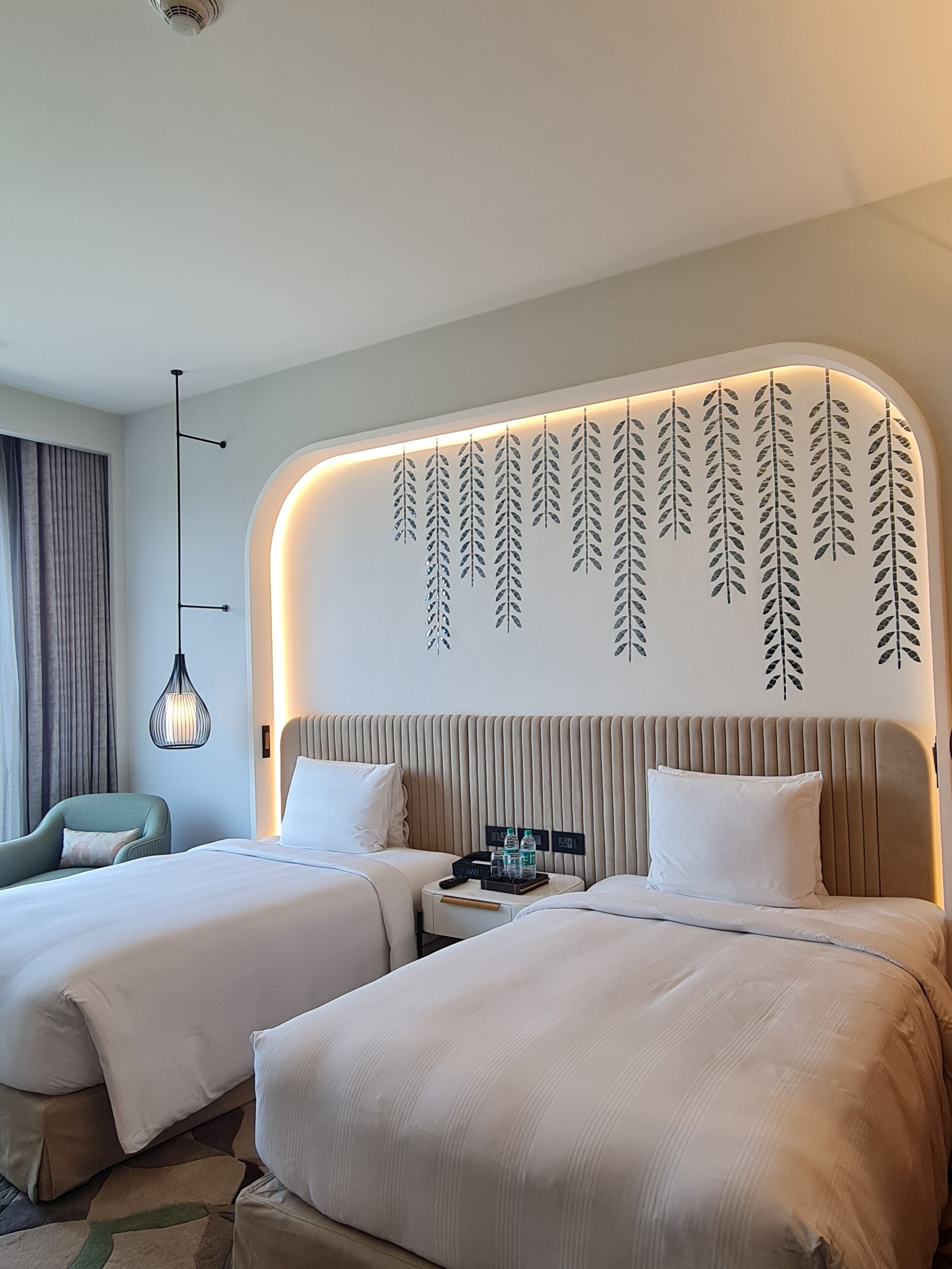 A hotel room with two beds, a bedside table with a lamp, and a wall with leaf patterns.