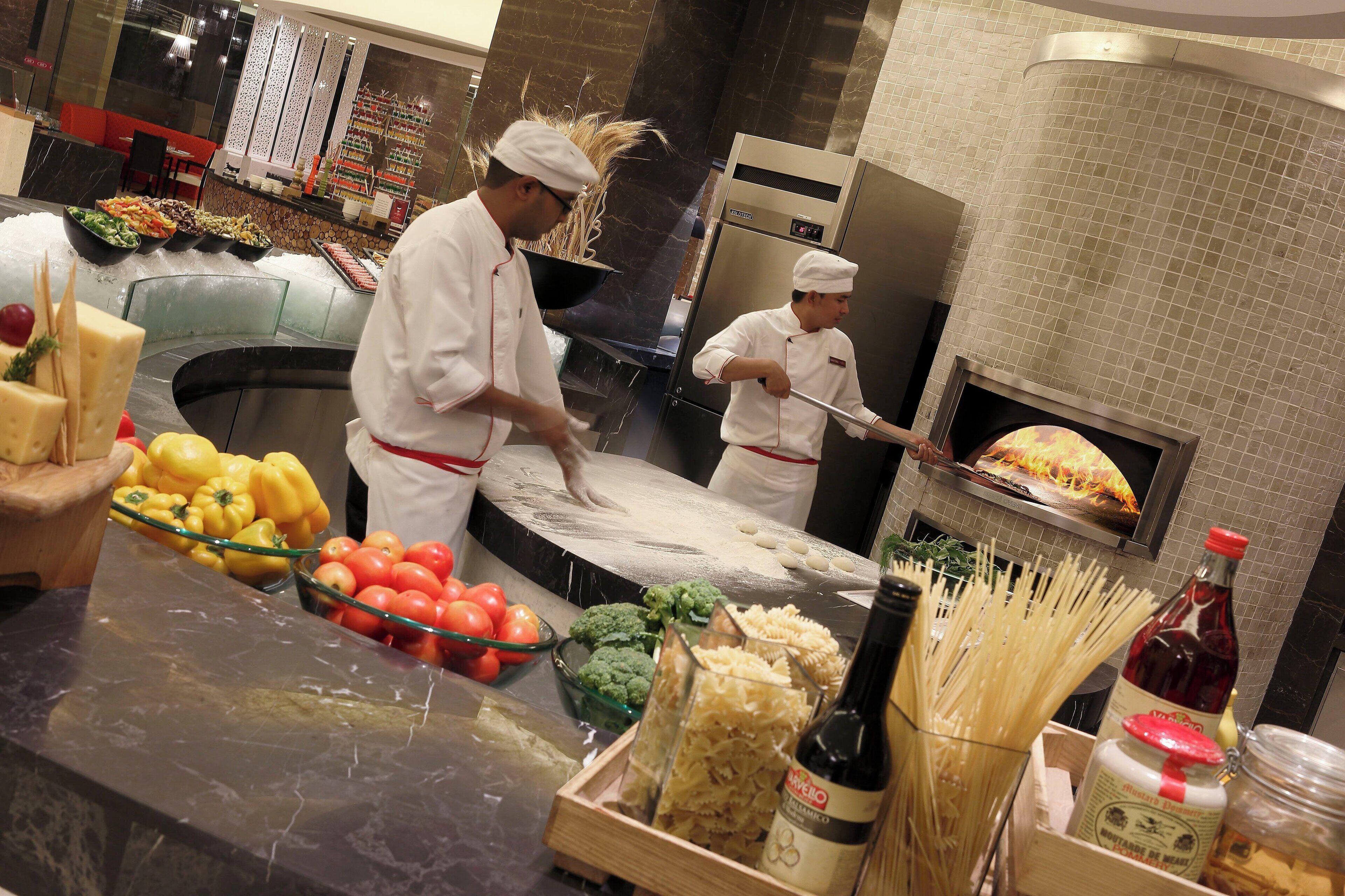 A kitchen area with chefs preparing food, a display of fresh produce, and a wood-fired oven.