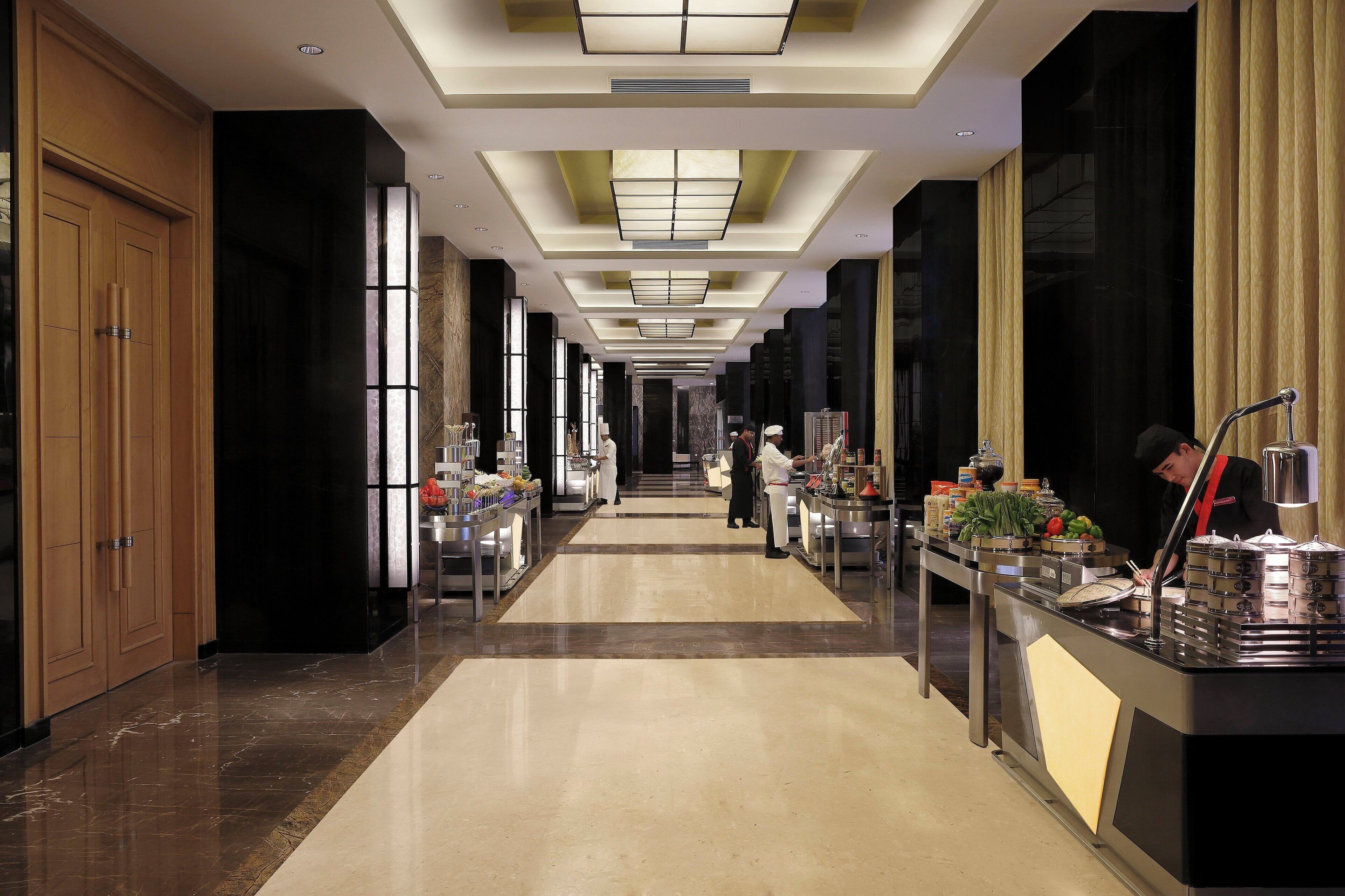 A hotel corridor with a long table displaying food items, a person serving, and a modern interior design.