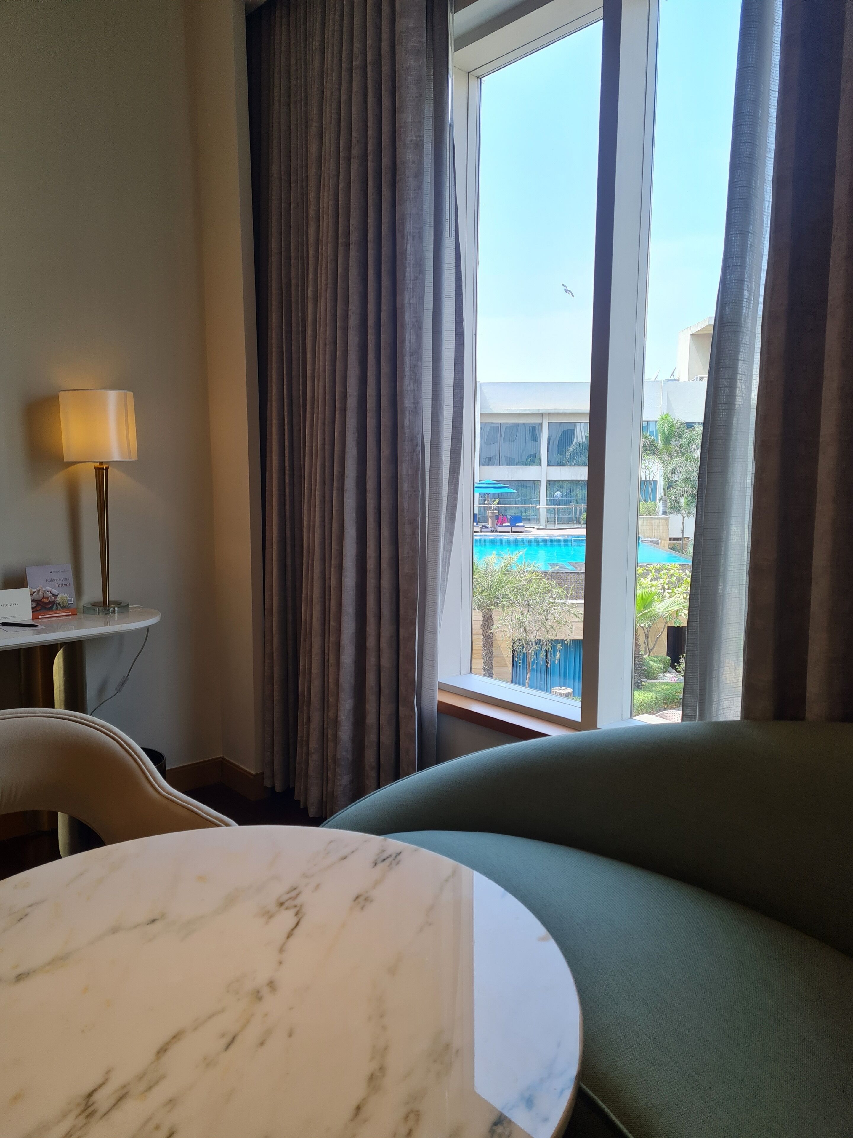 A hotel room with a view of a swimming pool, a sofa, a table with a marble top, and a lamp on a side table.