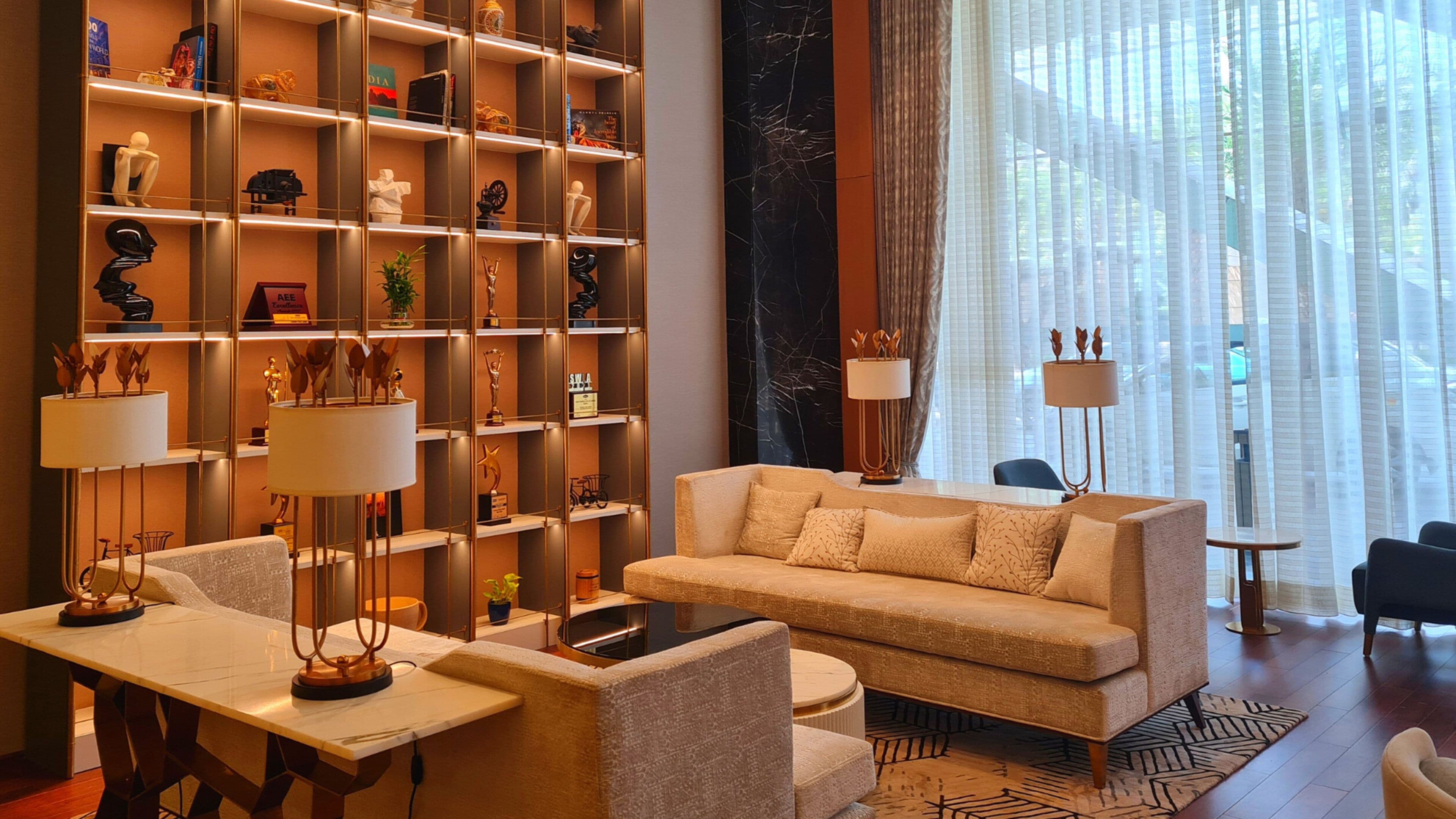 A modern living room with a sofa, armchairs, and a coffee table. There is a bookshelf filled with various decorative items and trophies. A large window with sheer curtains is visible in the background.