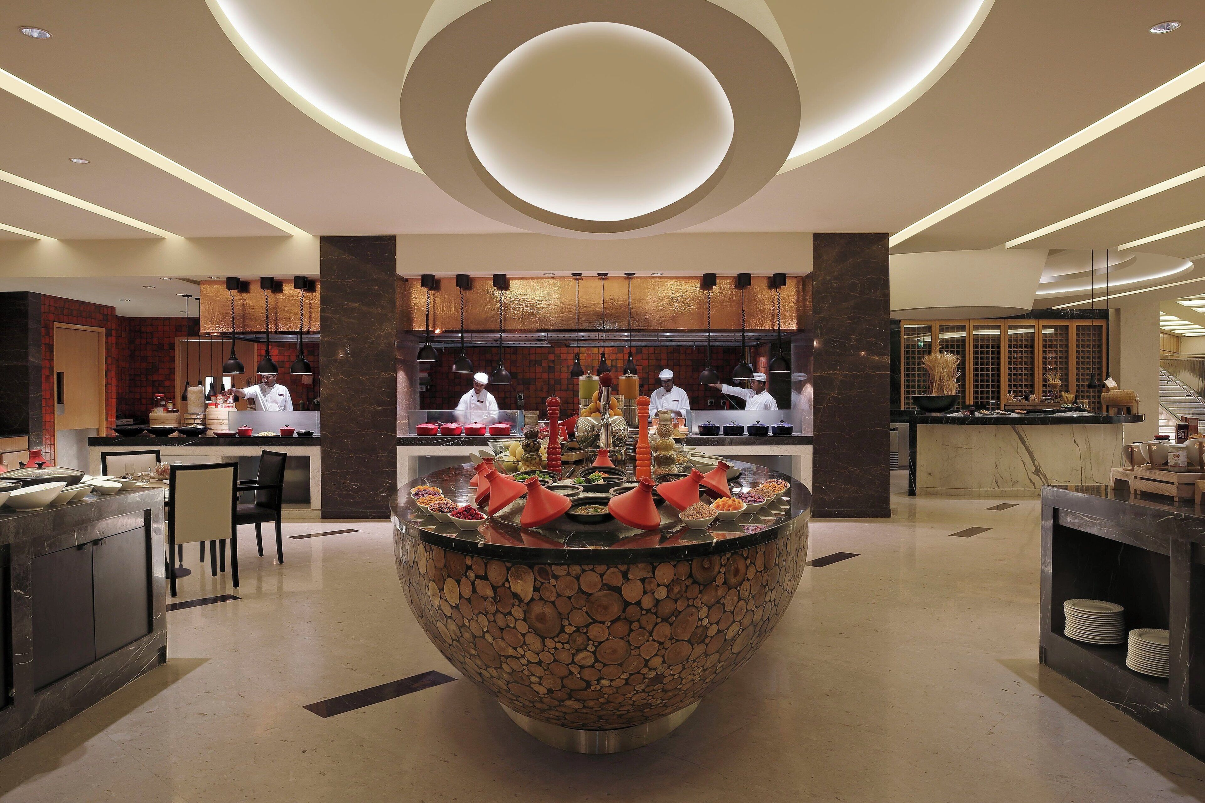Un moderno vestíbulo de hotel con una isla central que presenta un cuenco decorativo, un área de estar con sillas y una zona de cocina con chefs.