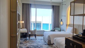 Ein Hotelzimmer mit einem großen Bett, einem kleineren Bett, einem Schreibtisch mit Stuhl, einem Schminktisch und einem großen Fenster mit Vorhängen.