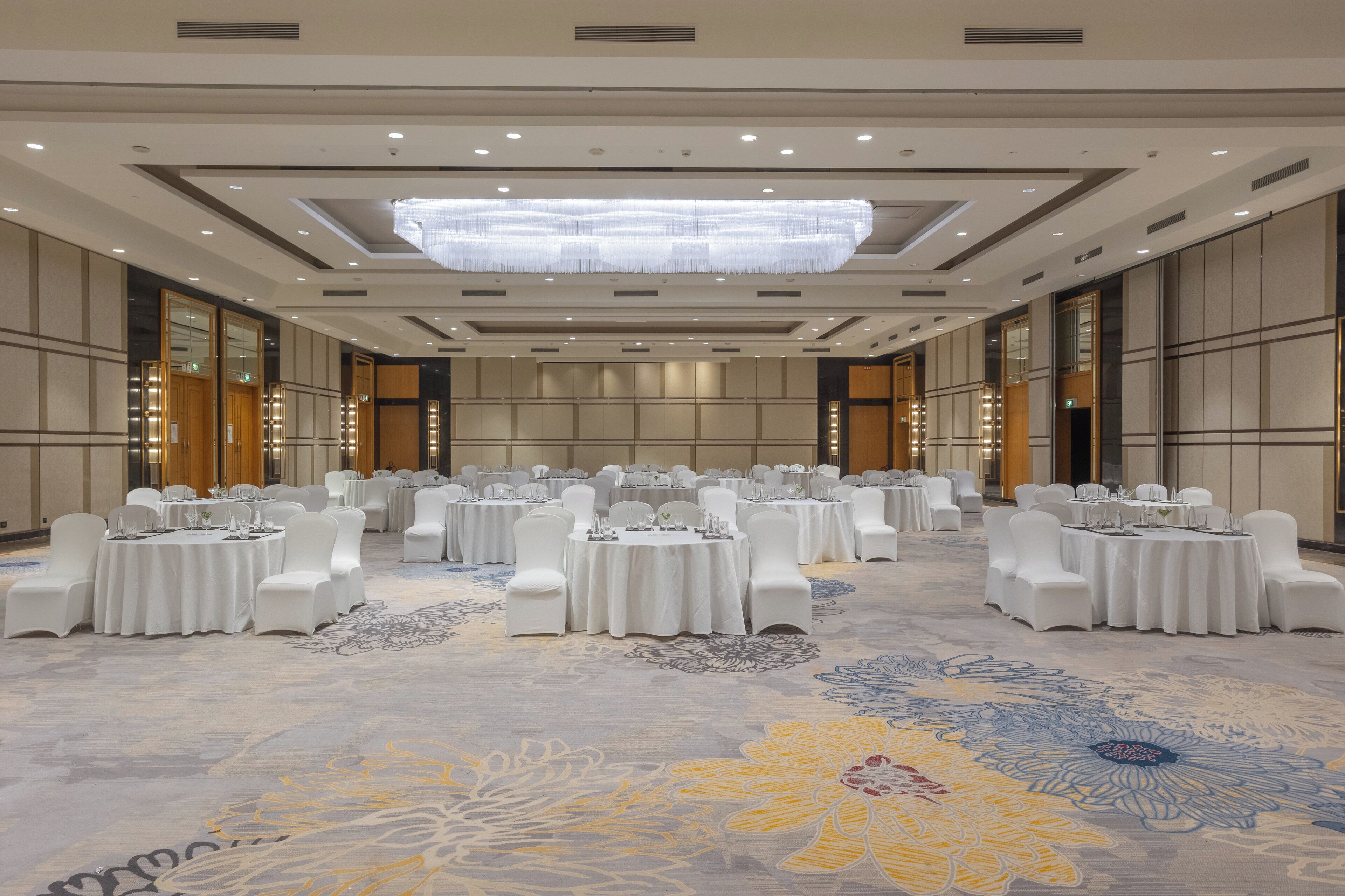 A large banquet hall with round tables set for dining, white chairs, and a decorative carpet.