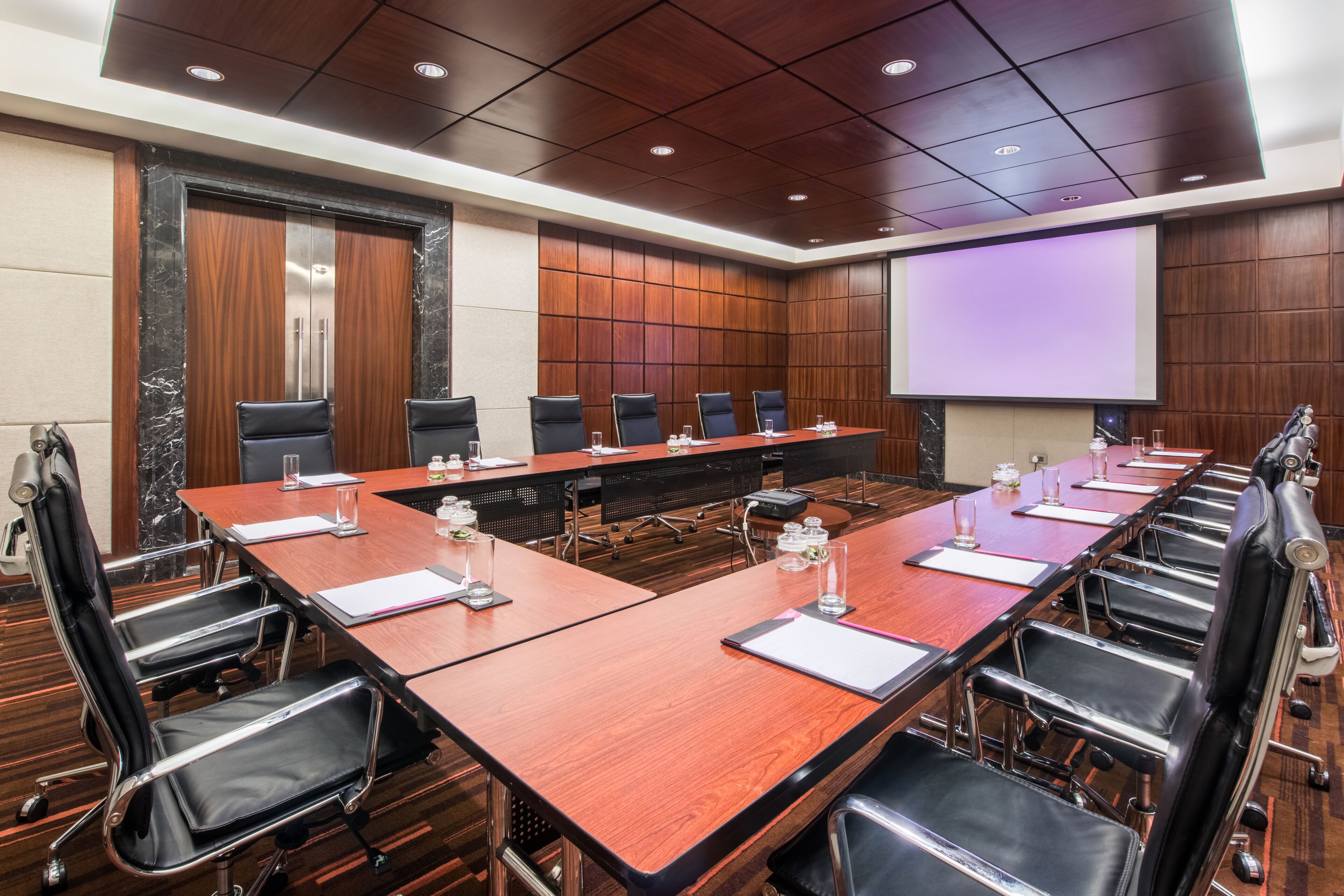 A conference room with a long table, multiple chairs, a projector screen, and a wooden panel wall.